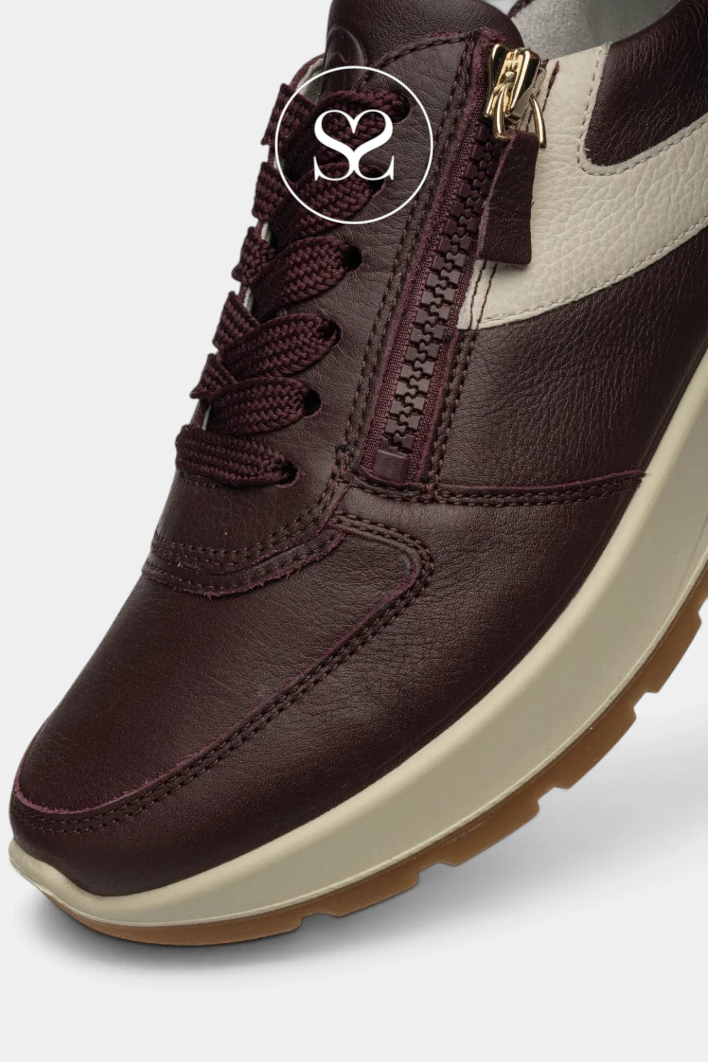 A close-up of a dark burgundy leather sneaker with a cream side panel, matching burgundy laces, a gold zipper pull, and a cream midsole on a light gray background.