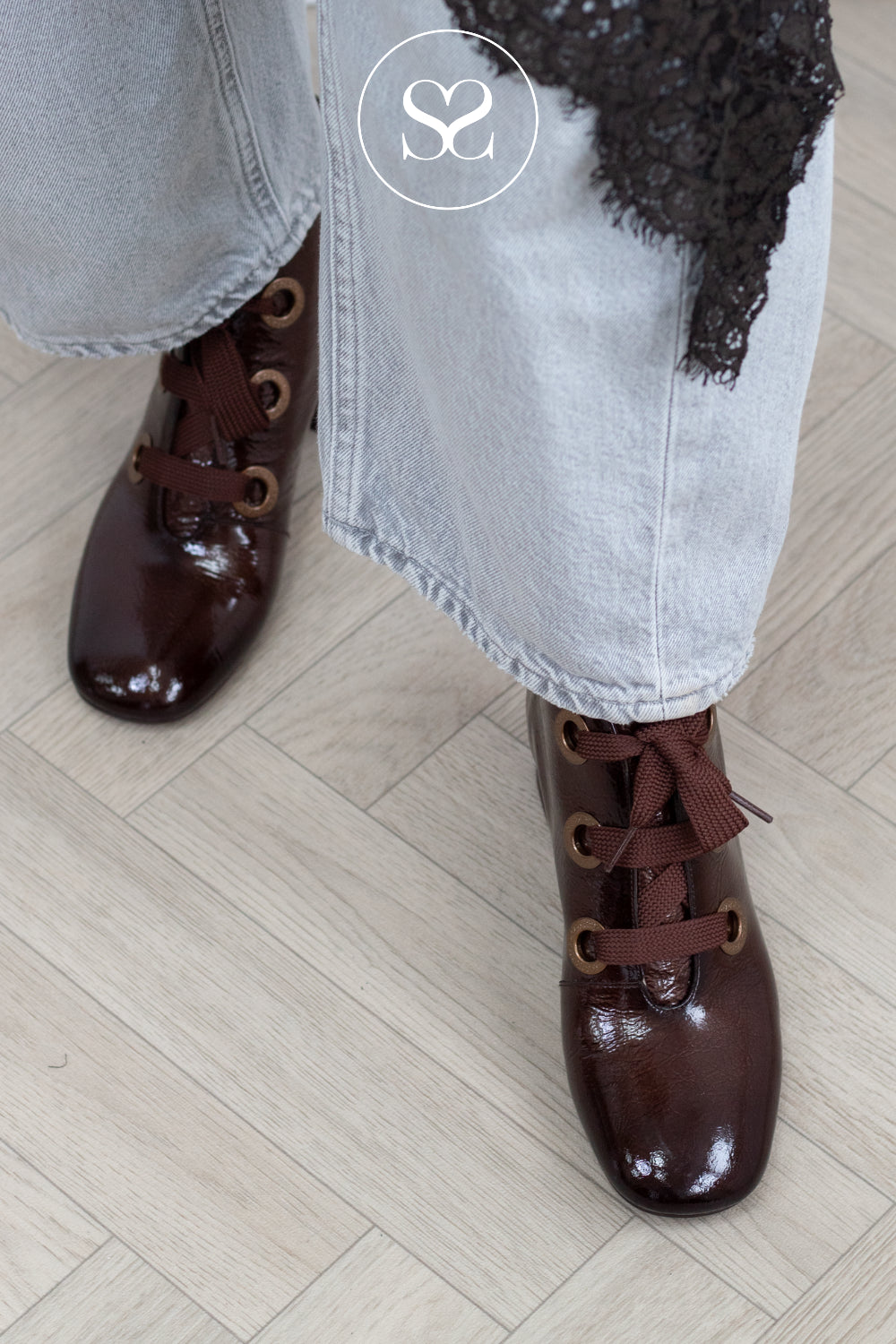 A close-up of a person wearing glossy dark brown lace-up ankle boots with metal eyelets, paired with light-wash jeans and a black lace top, standing on a light wood floor.