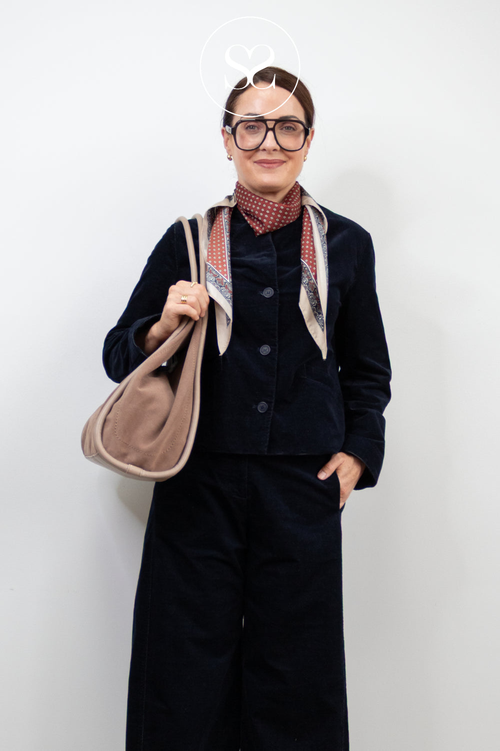 A woman wearing a dark navy corduroy jacket and matching wide-leg trousers, accessorized with a patterned scarf, glasses, and a Large Depeche beige shoulder bag, standing against a plain white background.