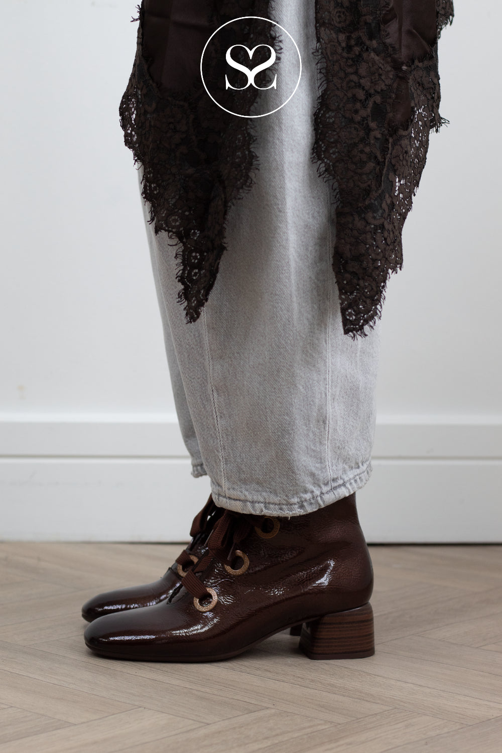 A person wearing dark brown patent leather lace-up ankle boots with a low block heel, styled with light gray jeans and a dark brown lace overlay, standing on a light wood floor.