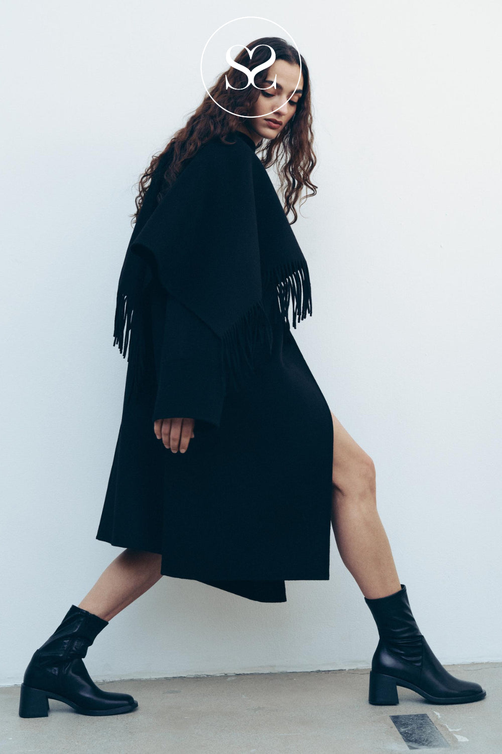 A woman models a black long-sleeve fringed coat with a high slit, paired with black Vagabond ankle boots, posing against a plain white wall.