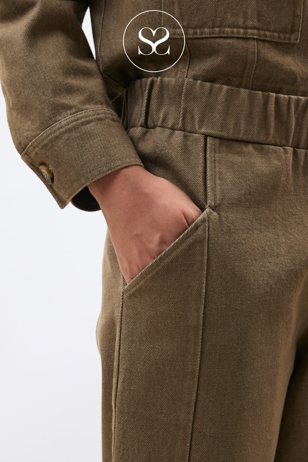 Close-up of a person wearing an olive brown jumpsuit, showing the side pocket with a hand inside and a circular 'SS' logo overlay near the top.