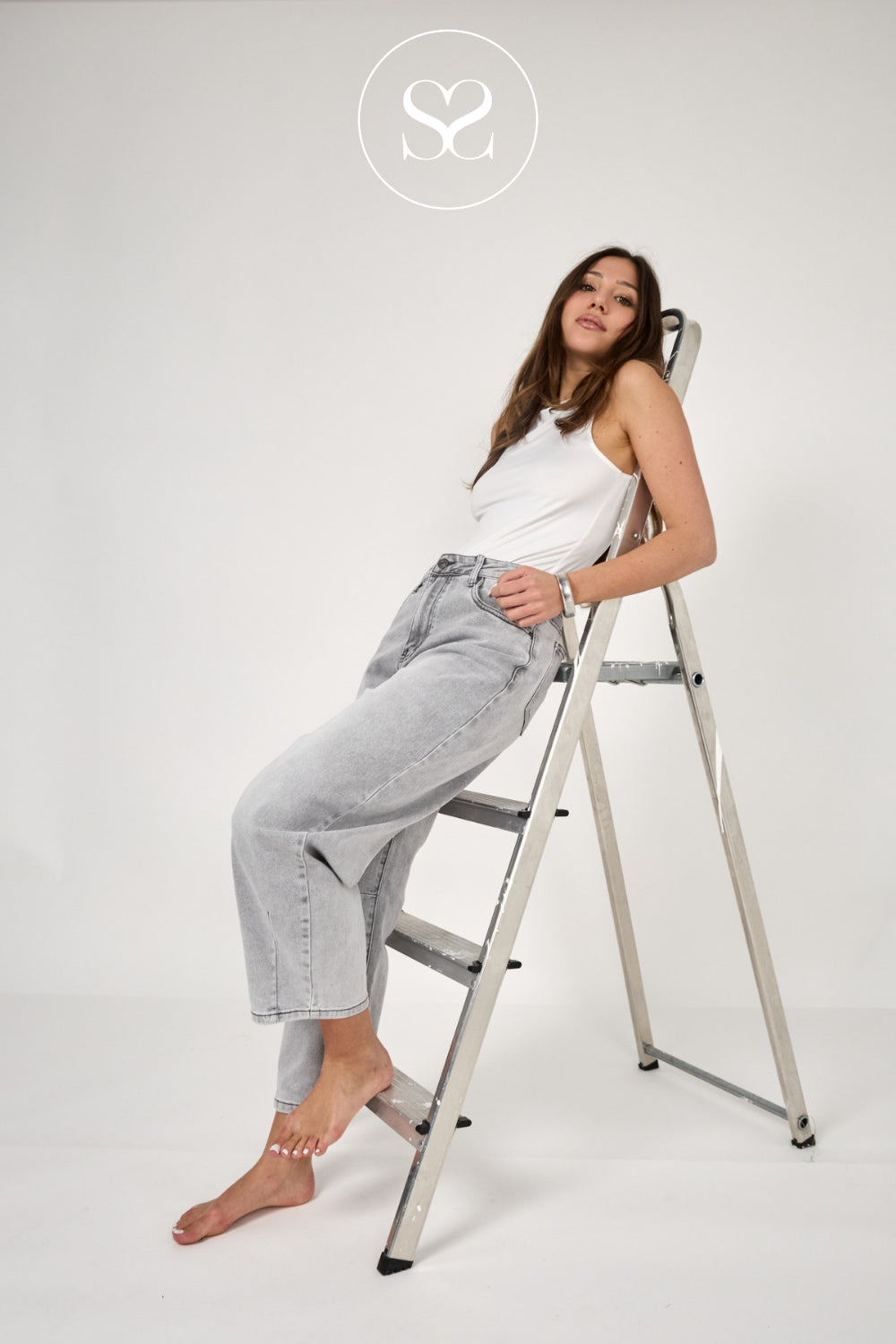 A woman posing on a silver metal step ladder against a plain white studio background, wearing a white tank top and light-wash wide-leg jeans. A circular logo with an 'S' is visible at the top center.