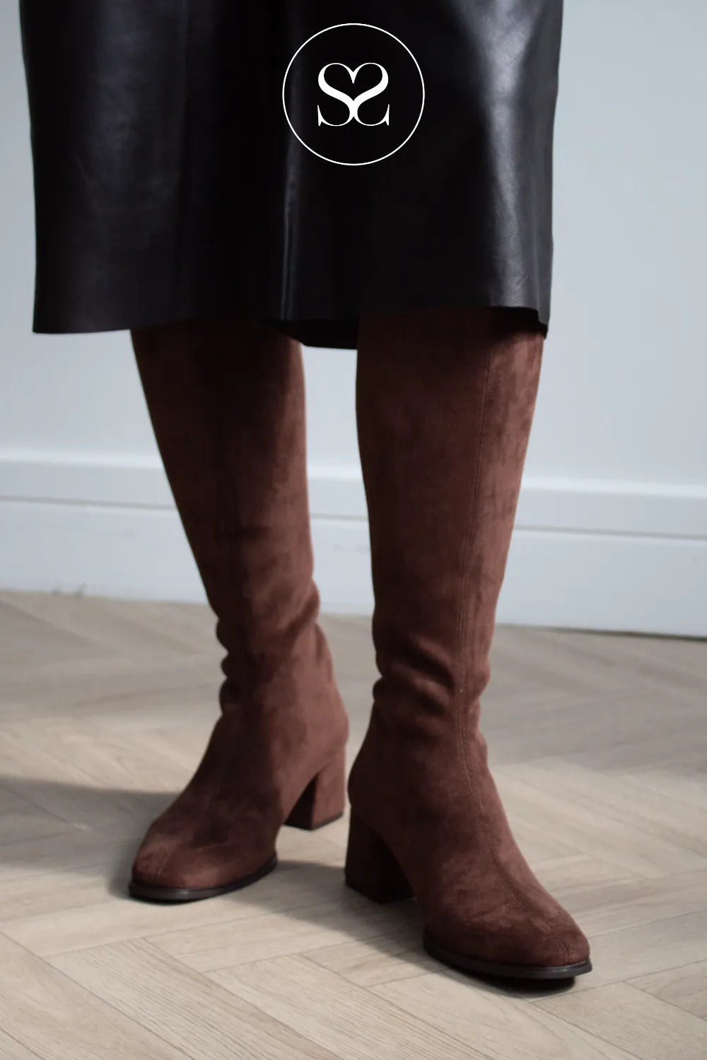 A person wearing dark brown suede knee boots with block heels, paired with a black skirt, standing on a light wood floor. Miss Elastic brown boots
