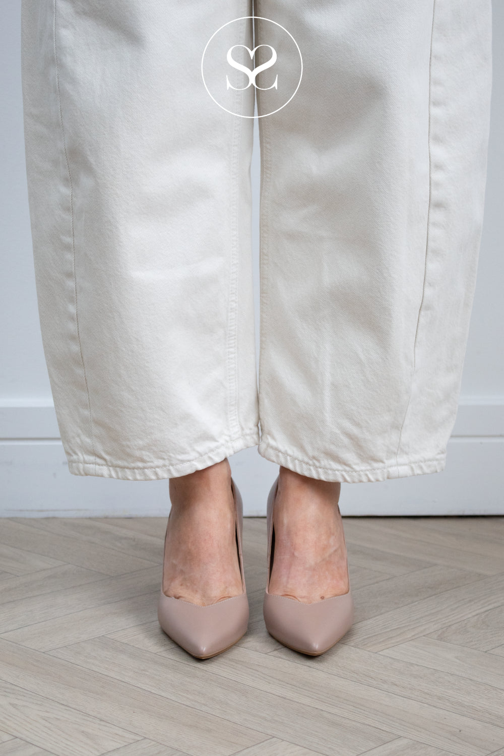 A person wearing off-white wide-leg trousers and taupe pointed-toe court high heeled shoes, standing on a light wood herringbone floor against a white wall. Lodi heels