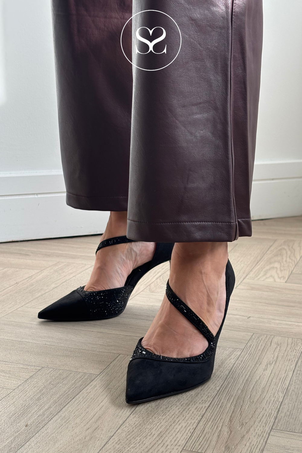 A person wearing black pointed-toe slingback heels with sparkling embellishments and crisscross ankle straps, paired with dark brown leather pants, standing on a light wood herringbone floor.