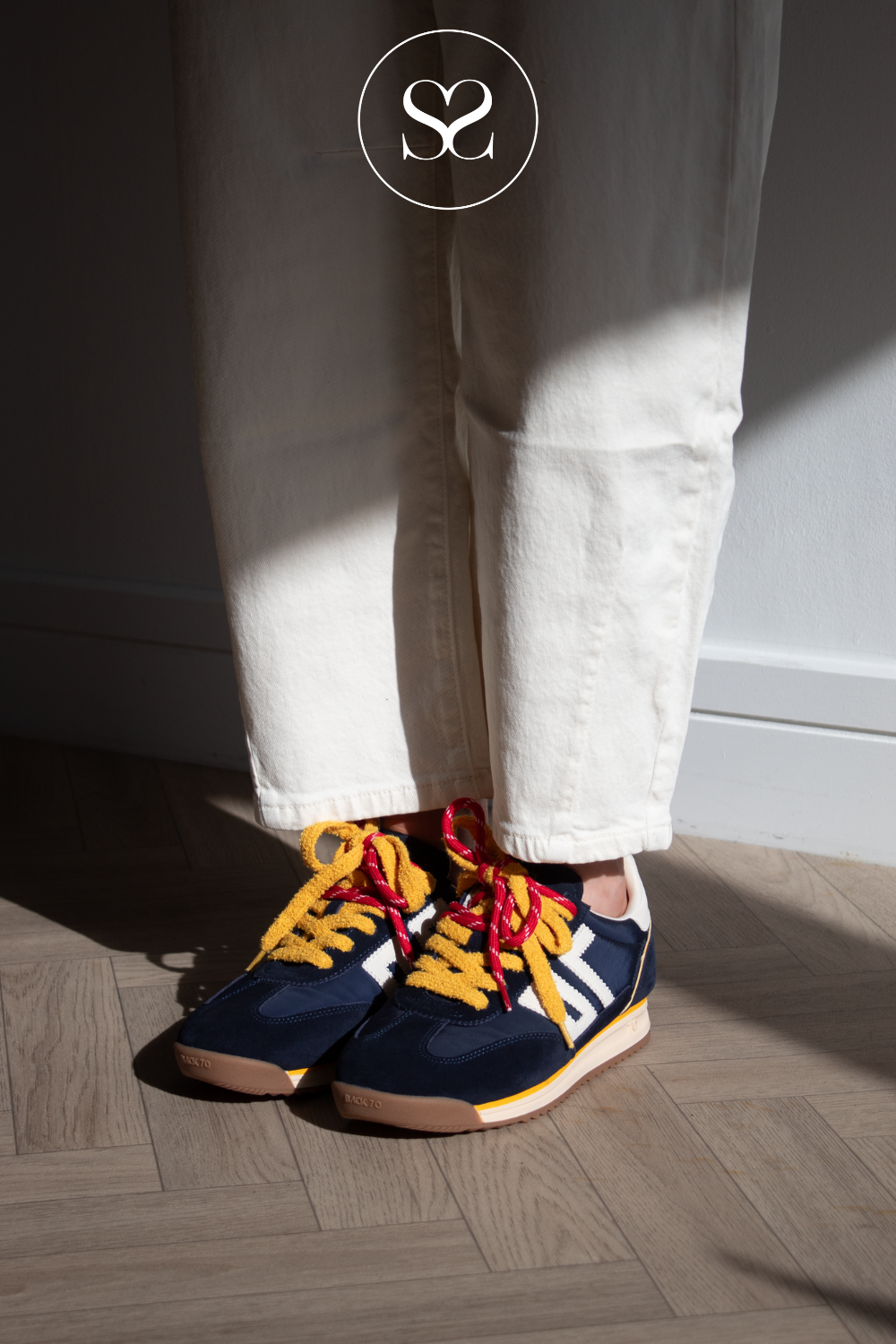 A person wearing cream-colored pants and navy blue sneakers with yellow and red laces, standing on a wooden floor in direct sunlight.