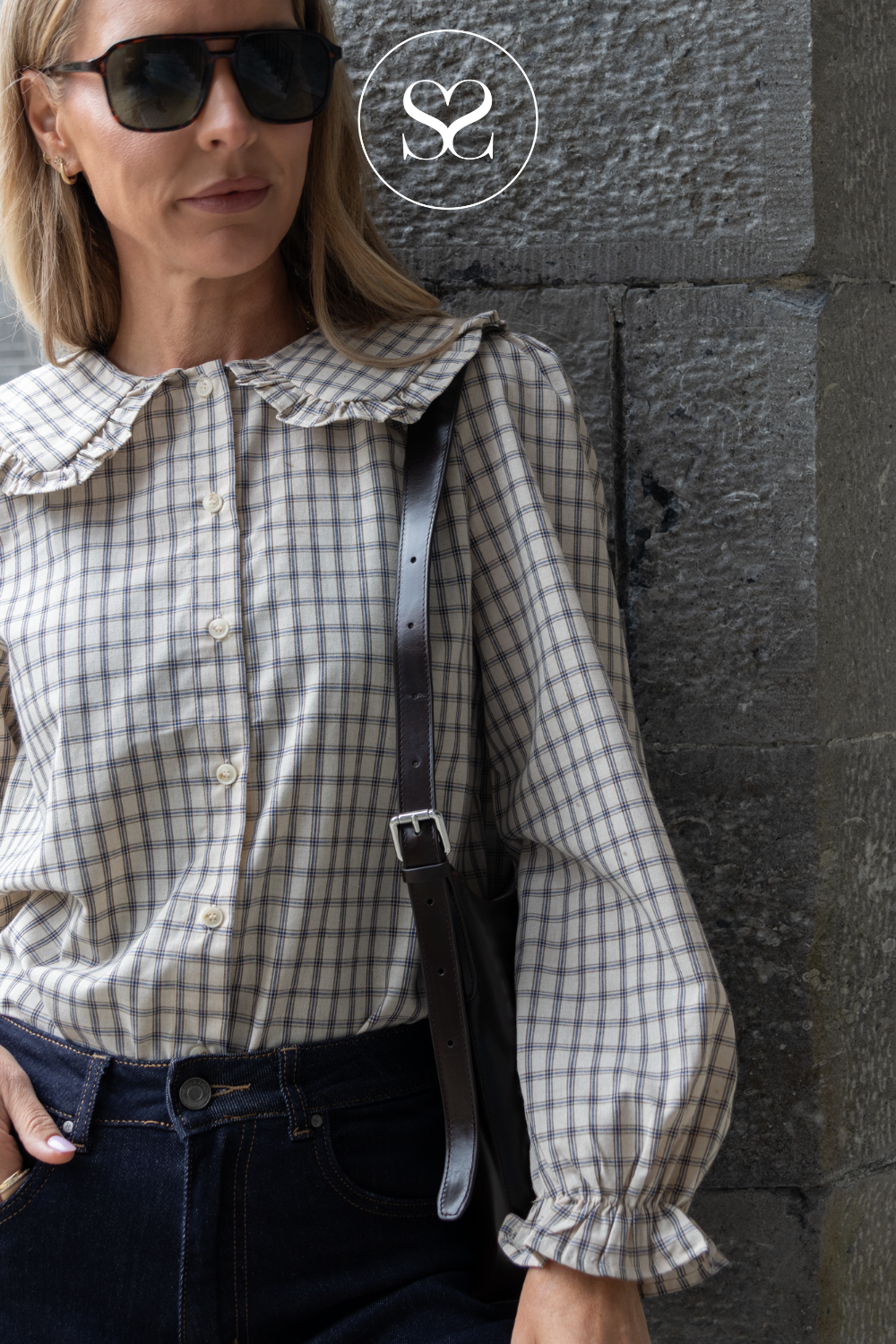 A woman wearing a beige and navy blue gingham long-sleeve blouse with a ruffled collar, dark sunglasses, and dark jeans, standing against a textured gray stone wall.