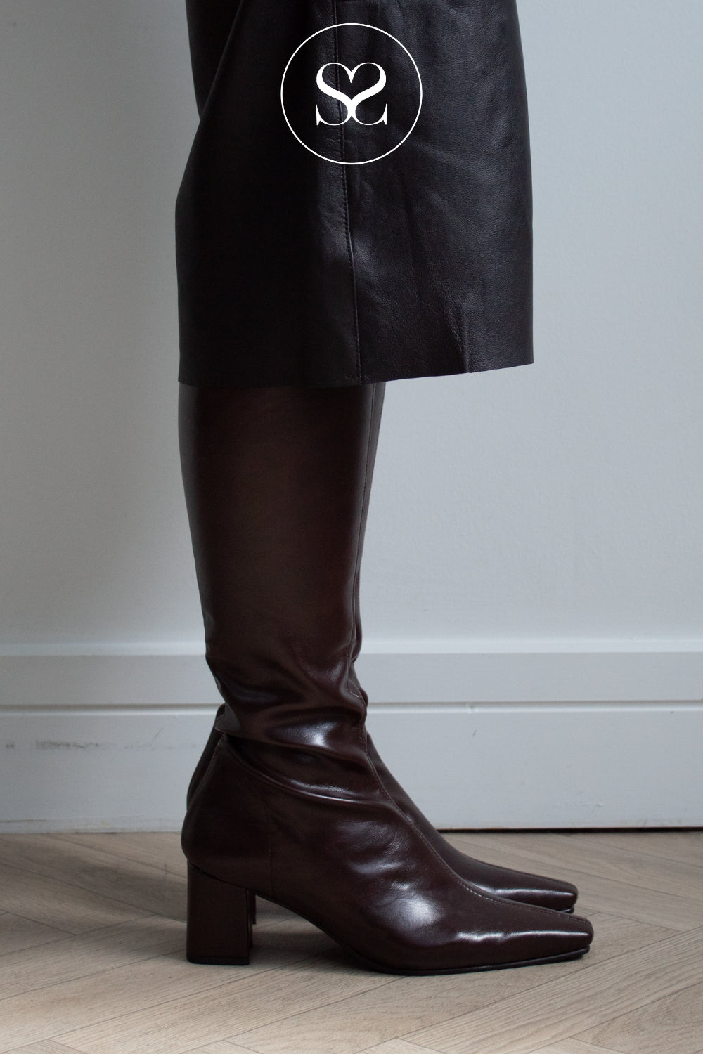 A close-up of a person's leg wearing a dark brown leather knee-high boot with a block heel, paired with a black skirt. A white circular logo with 'Sheneil Shoes' is visible on the skirt.