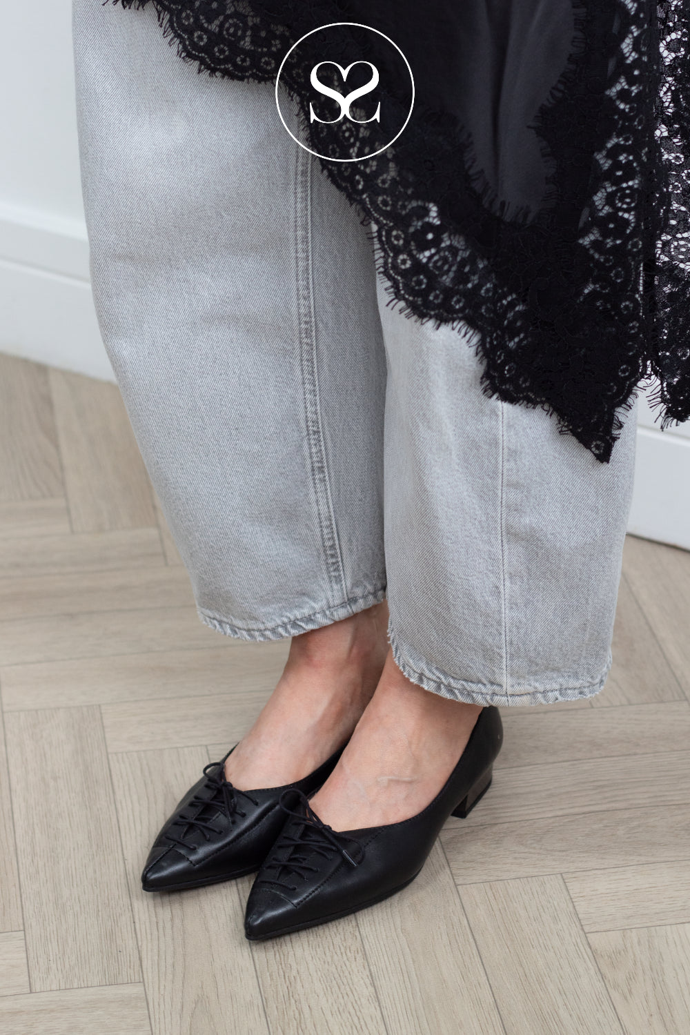 A close-up of a person wearing black pointed-toe flats with lace-up detailing, paired with light gray cropped jeans and a black lace-trim top, standing on a light wood herringbone floor.