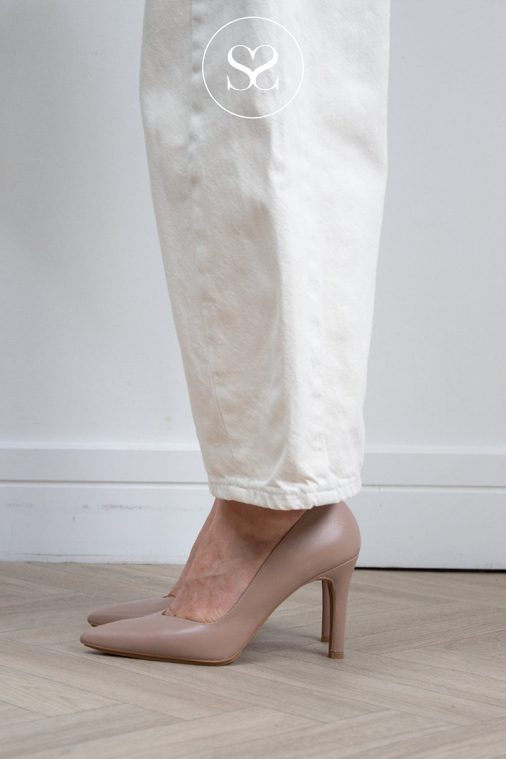 A person wearing beige pointed-toe high heels with white trousers, standing on a light wood herringbone floor against a white wall. Lodi Taupe rabot shoes in Ireland