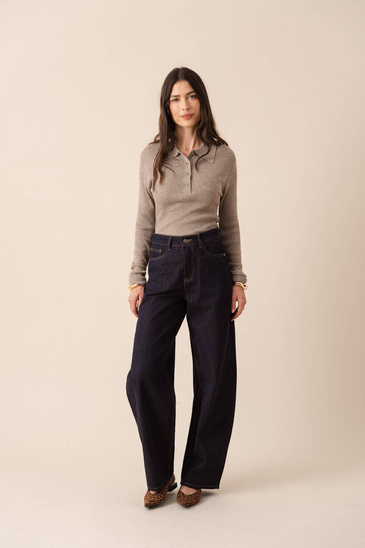 A woman modeling a light taupe knit polo shirt and dark navy wide-leg jeans against a solid beige studio background.