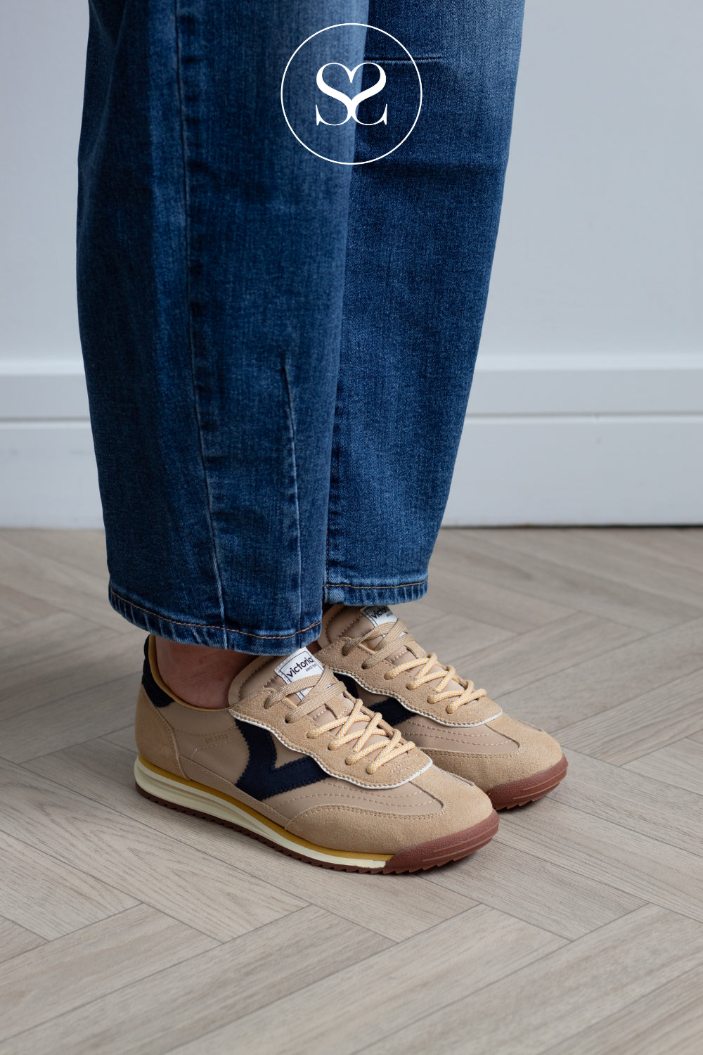 A person wearing beige suede and leather low-top sneakers with dark navy accents, paired with blue jeans, standing on a light wood herringbone floor.