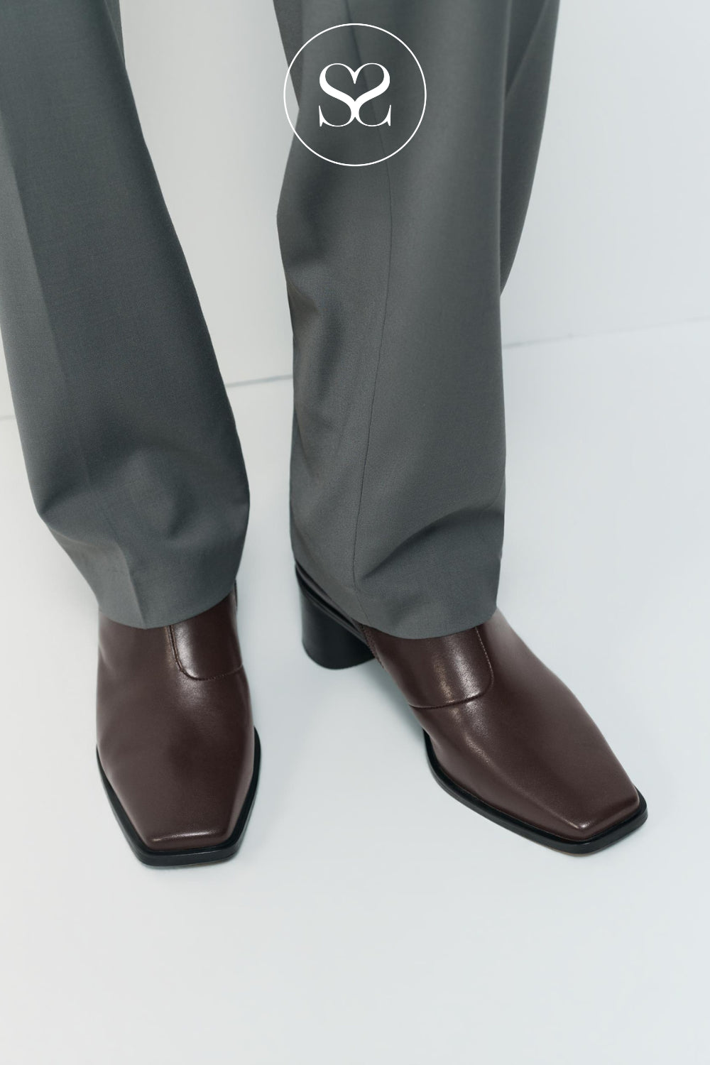 A close-up of a woman's legs wearing gray trousers and dark brown leather square-toe ankle boots with low block heels from Vagabond, shown against a clean white studio background.