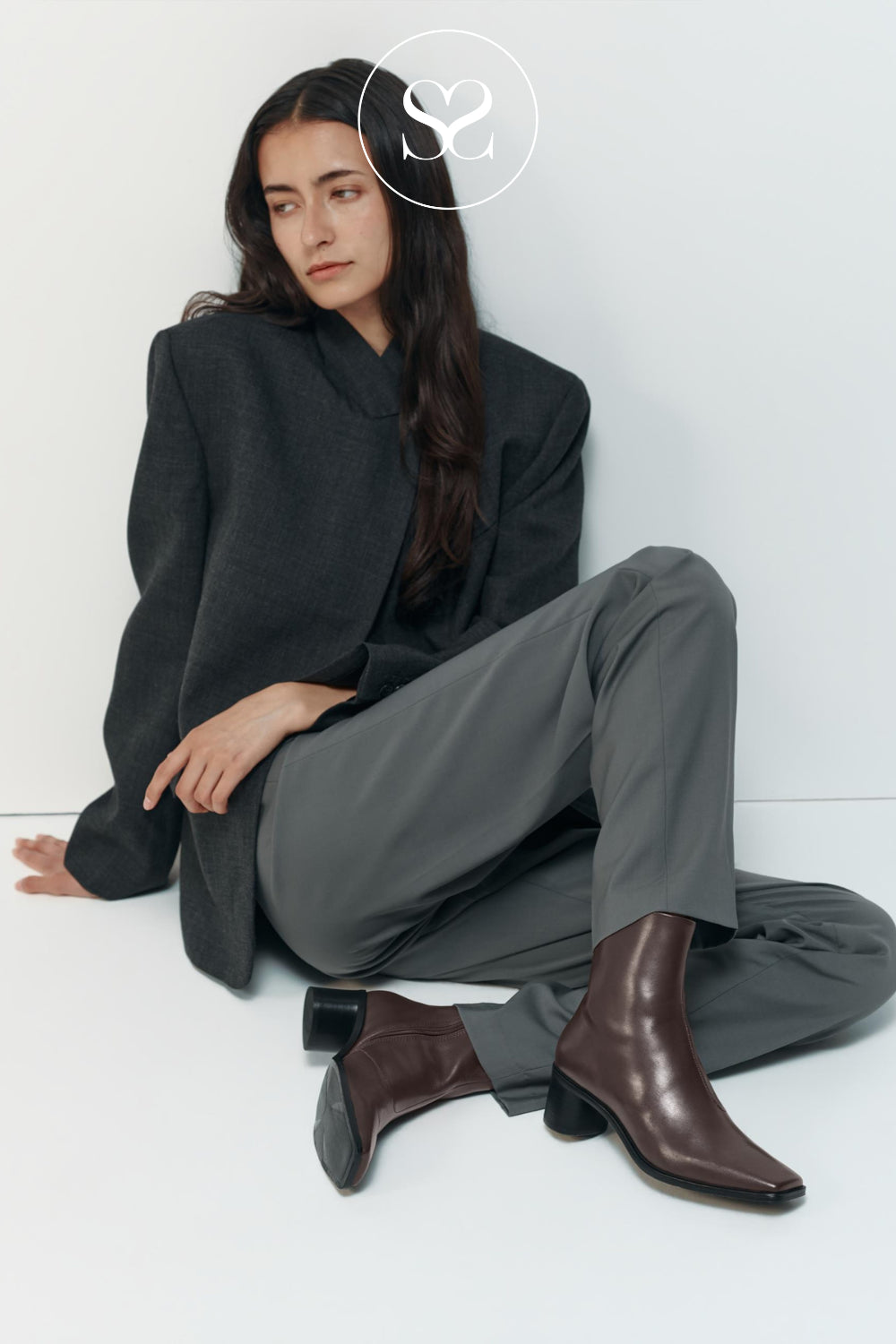 A model sits on the floor wearing a dark oversized blazer, gray trousers, and dark brown pointed-toe ankle boots (Vagabond Fay) against a clean white studio background in Ireland.