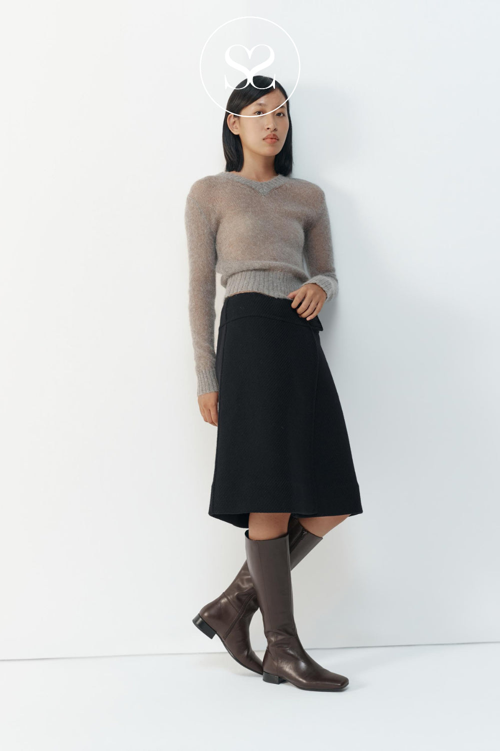 A model stands against a white studio wall wearing a taupe knit sweater, a black midi skirt, and brown knee-high boots from Vagabond. A small white circular logo is visible above the model's head.