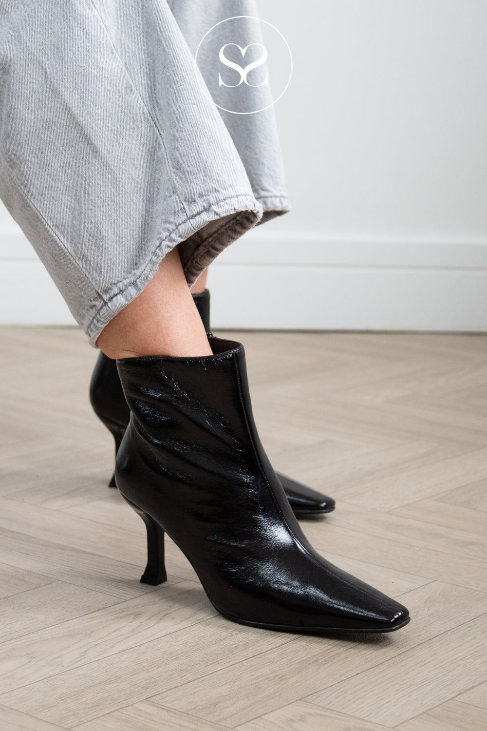 A person wearing Lodi high shine black pointed-toe ankle boots with kitten heels, paired with light gray cropped jeans, standing on a light wood floor against a white wall.