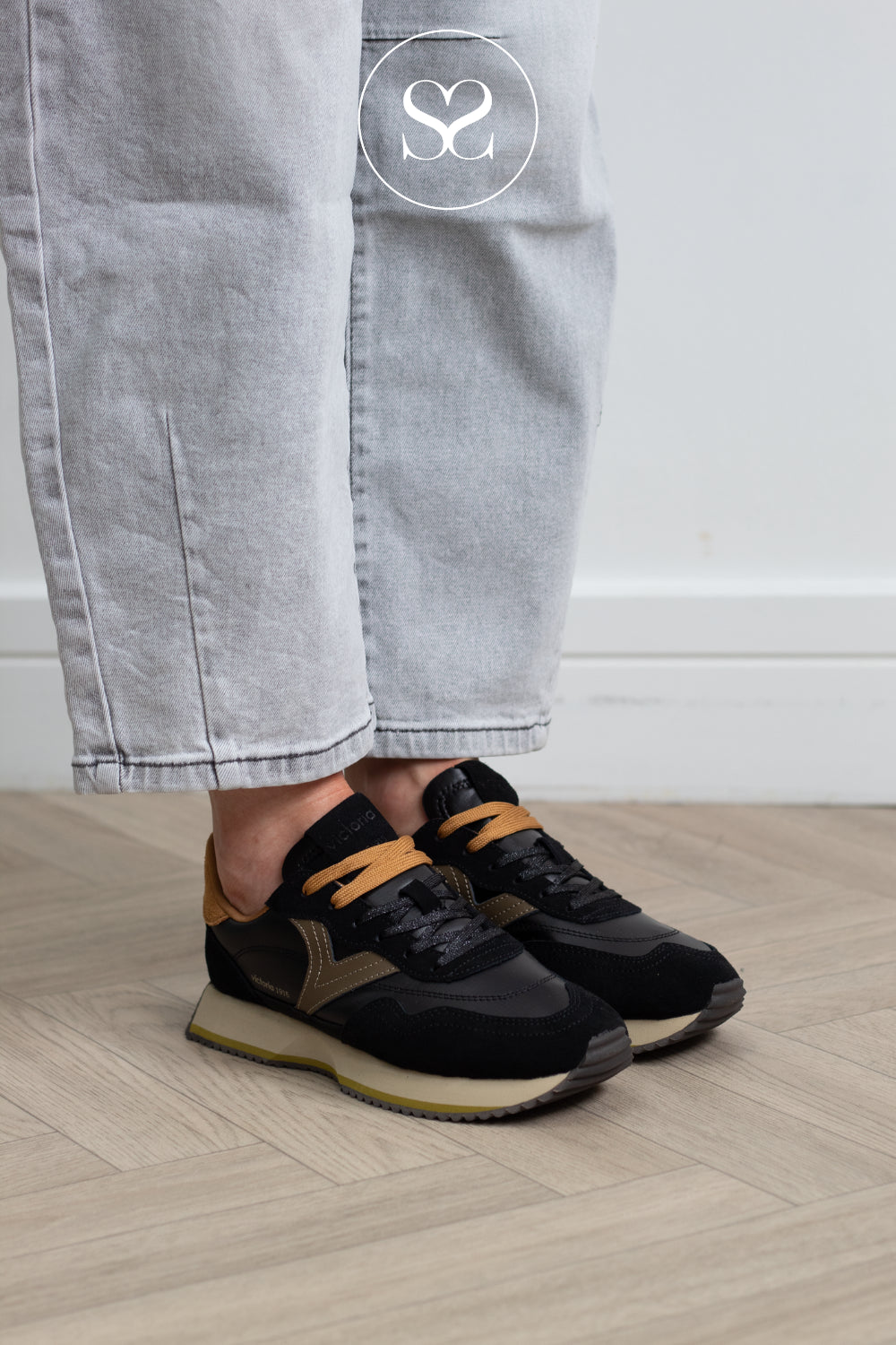A person wearing black and tan athletic sneakers with light gray jeans, standing on a light wood herringbone floor against a white wall.