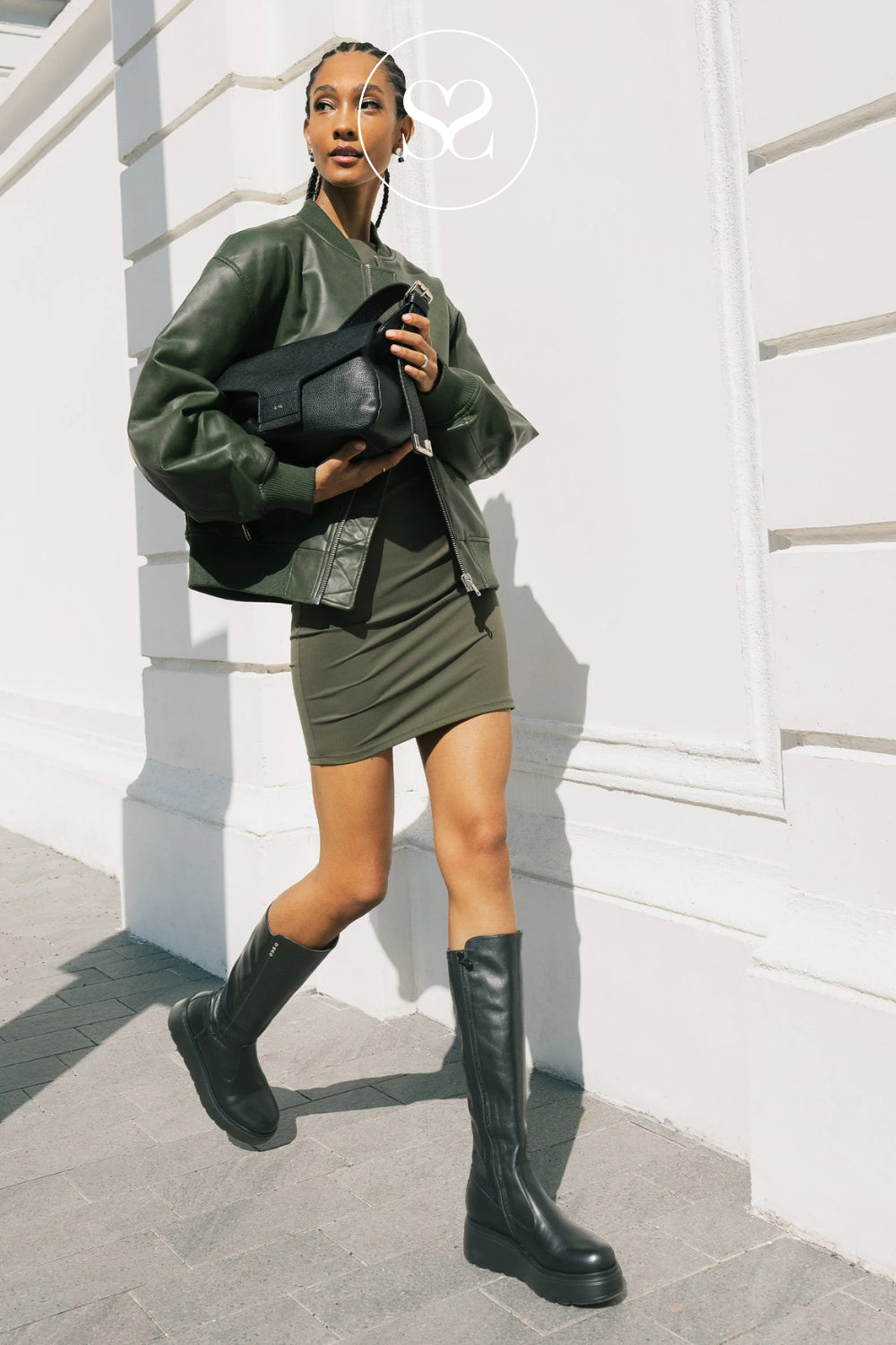 A woman models an olive green leather jacket and matching mini skirt set, paired with Nero Giardini black leather knee-high boots and a black shoulder bag, standing against a white building wall in Ireland