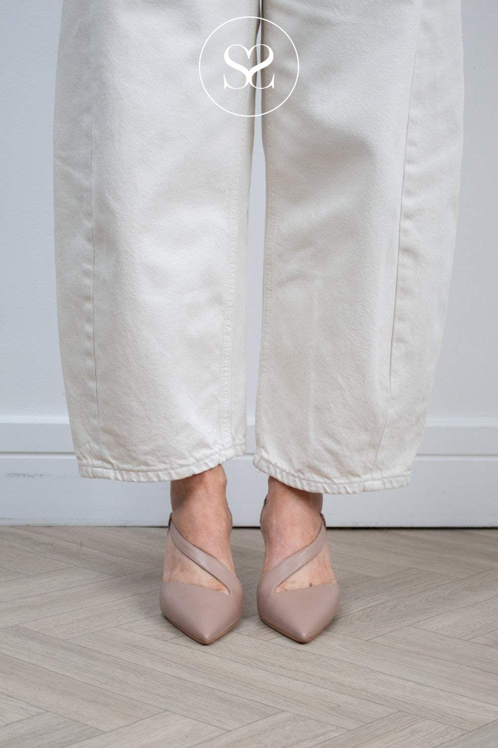 A Woman wearing off-white wide-leg trousers and taupe pointed-toe slingback heels, standing on a light wood herringbone floor against a white wall. Lodi nude shoes in Ireland