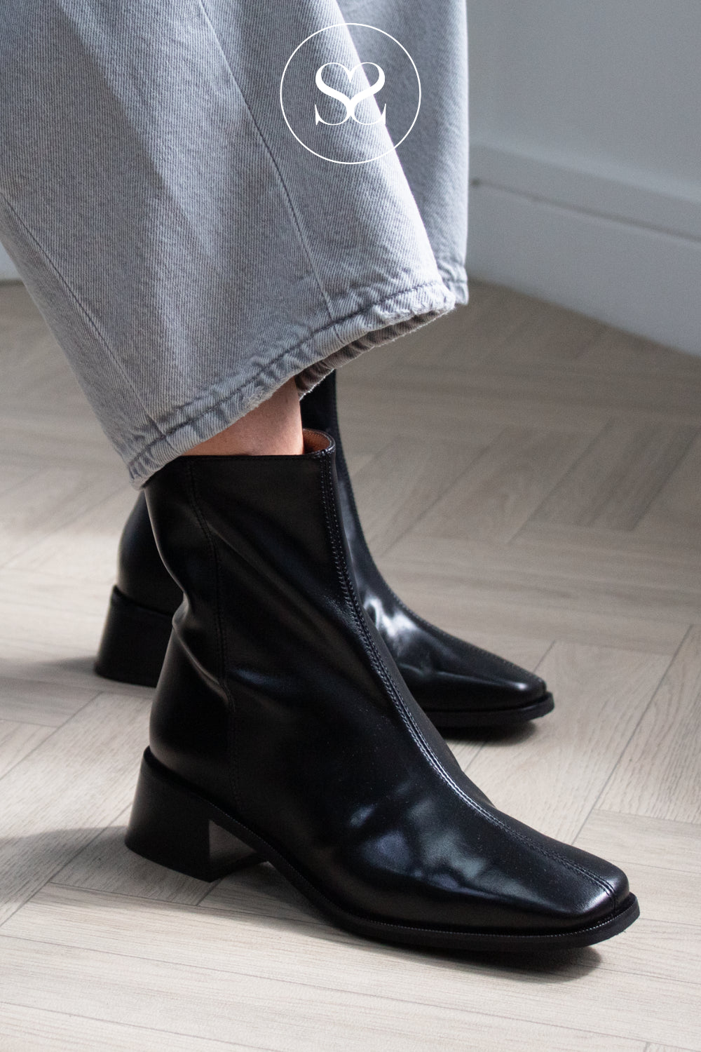 A person wearing glossy black ankle boots with low block heels and square toes from Wonders in Ireland, paired with light gray jeans, standing on a light wood floor.