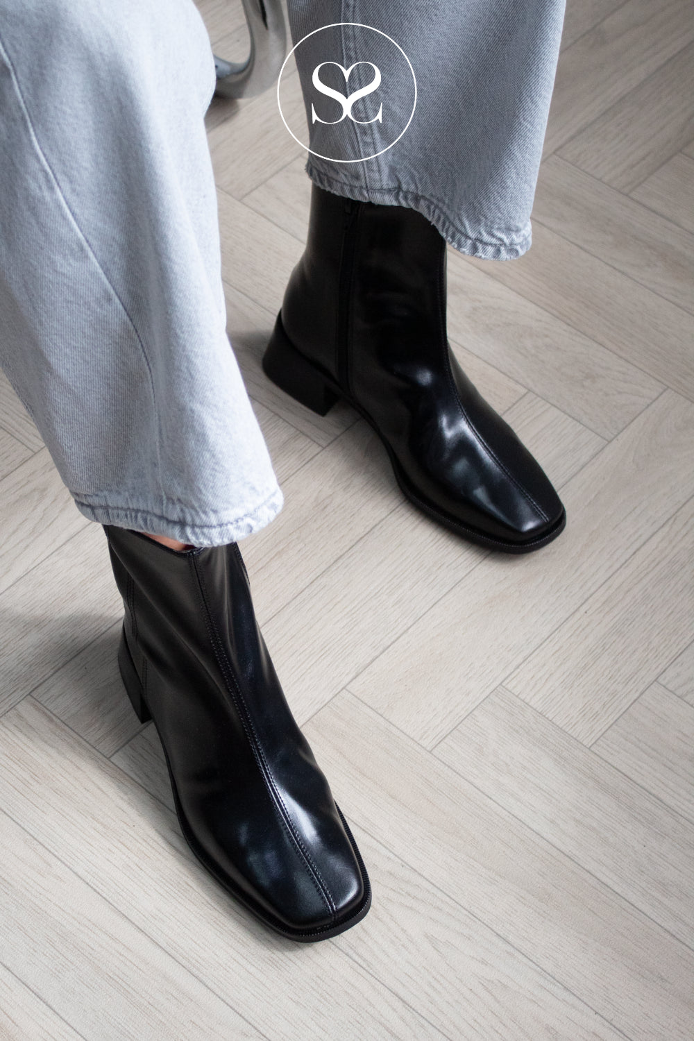 A person wearing High Shine black leather ankle boots with a square toe and low block heel, paired with light-wash blue jeans, standing on a light wood herringbone floor.