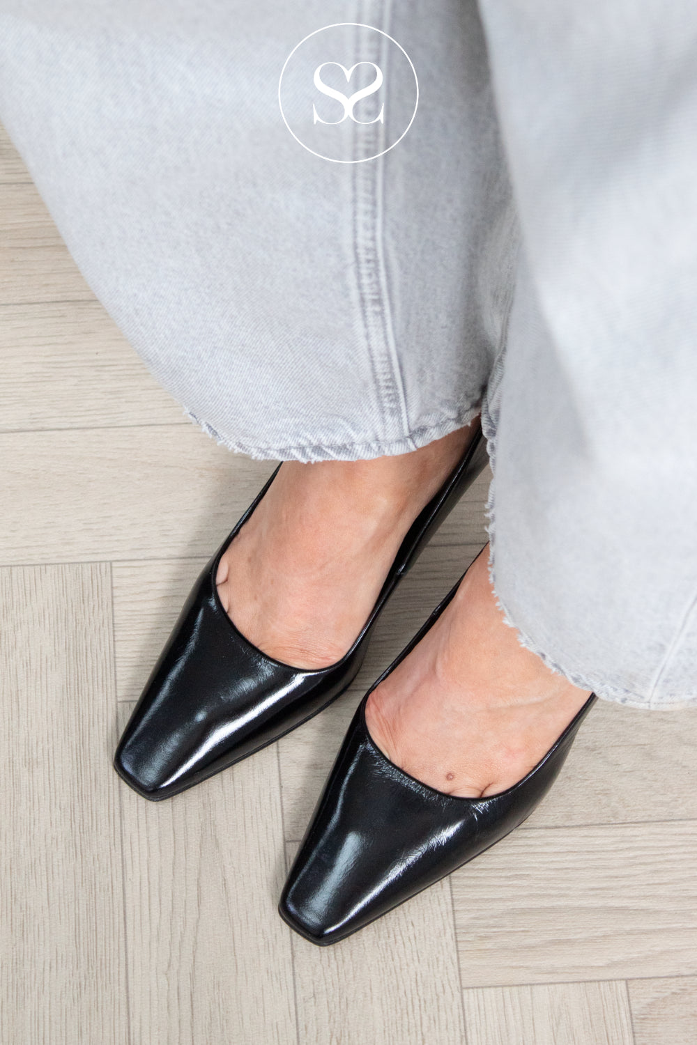 A close-up of a person wearing shiny black leather pointed-toe kitten heels with light grey jeans, standing on a light wood floor. A circular logo watermark for sheneil shoes is visible near the top center. shoes are lodi pauline heels