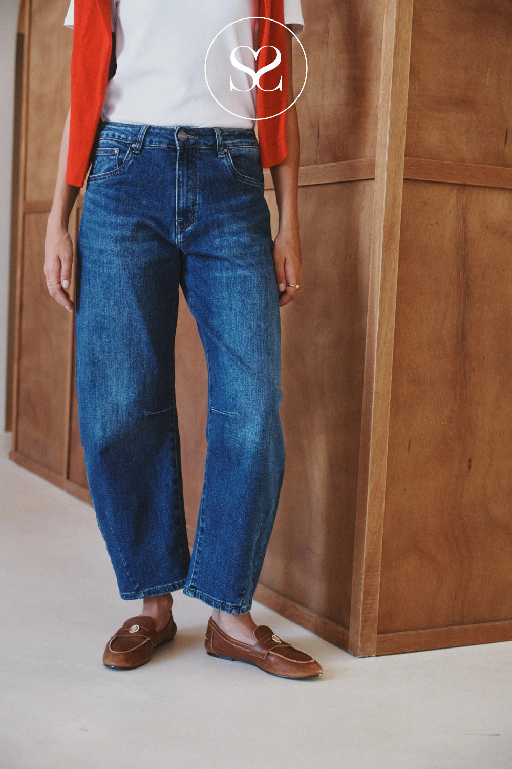 A person wearing high-waisted, barel leg dark blue denim jeans with brown leather loafers, standing in front of a wooden paneled wall. A white t-shirt and red cardigan are partially visible, and a circular 's2' logo watermark is overlaid at the top.