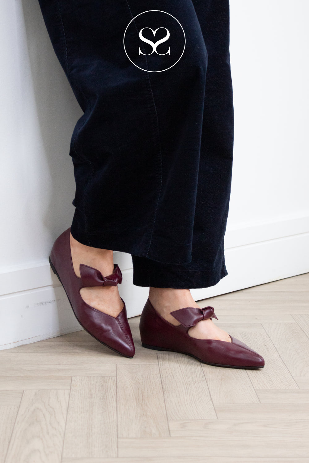 A person wearing dark navy corduroy trousers and burgundy pointed-toe flat shoes with bow details, standing on a light wood herringbone floor against a white wall.
