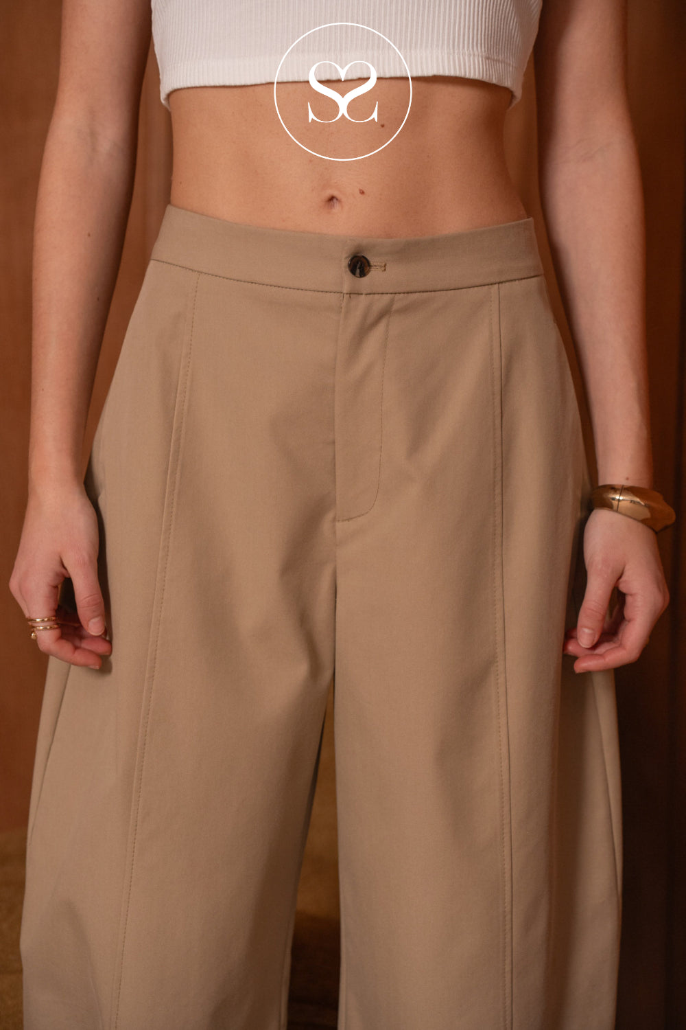 A close-up of a model wearing high-waisted, wide-leg taupe pants with a single button closure at the front waistband, paired with a white ribbed crop top against a warm brown background.
