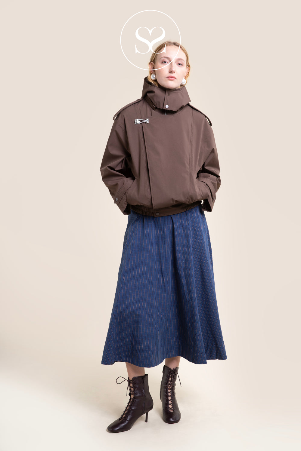 A model stands against a plain beige studio background wearing a brown cropped jacket with a high collar from Flotte, a blue pleated midi skirt, and dark brown lace-up ankle boots. A white circular Sheneil Shoes logo is visible above the model's head.
