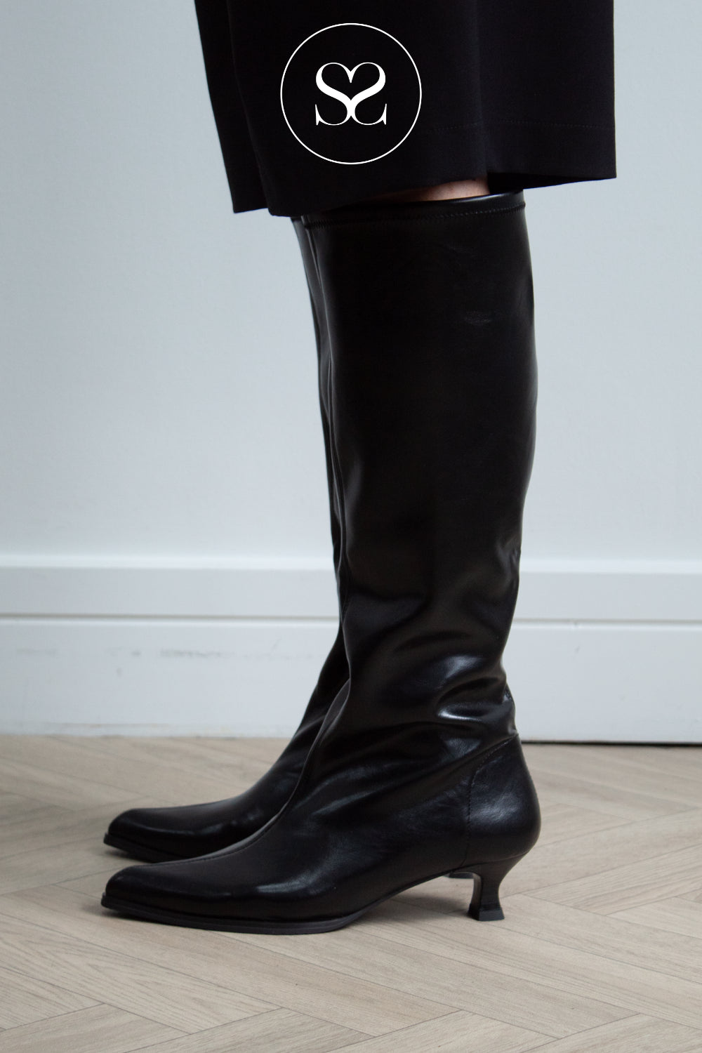 A close-up of a woman's lower legs wearing black leather look slouch knee-high boots with kitten heels, paired with black trousers featuring a white circular sheneil shoes logo overlay.