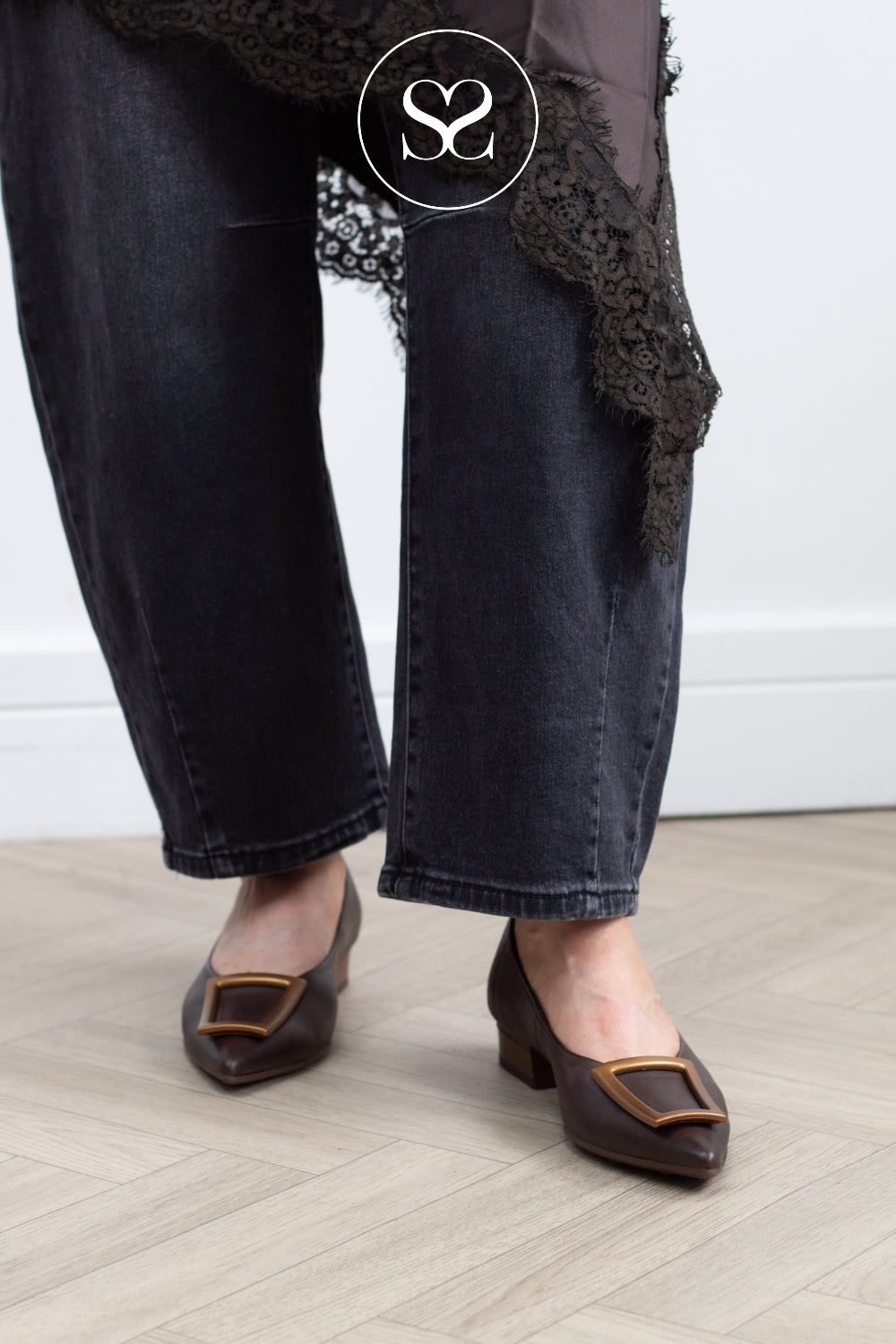 A person wearing dark charcoal wide-leg jeans with black lace trim and brown pointed-toe flats with gold square hardware, standing on a light wood floor against a white wall.