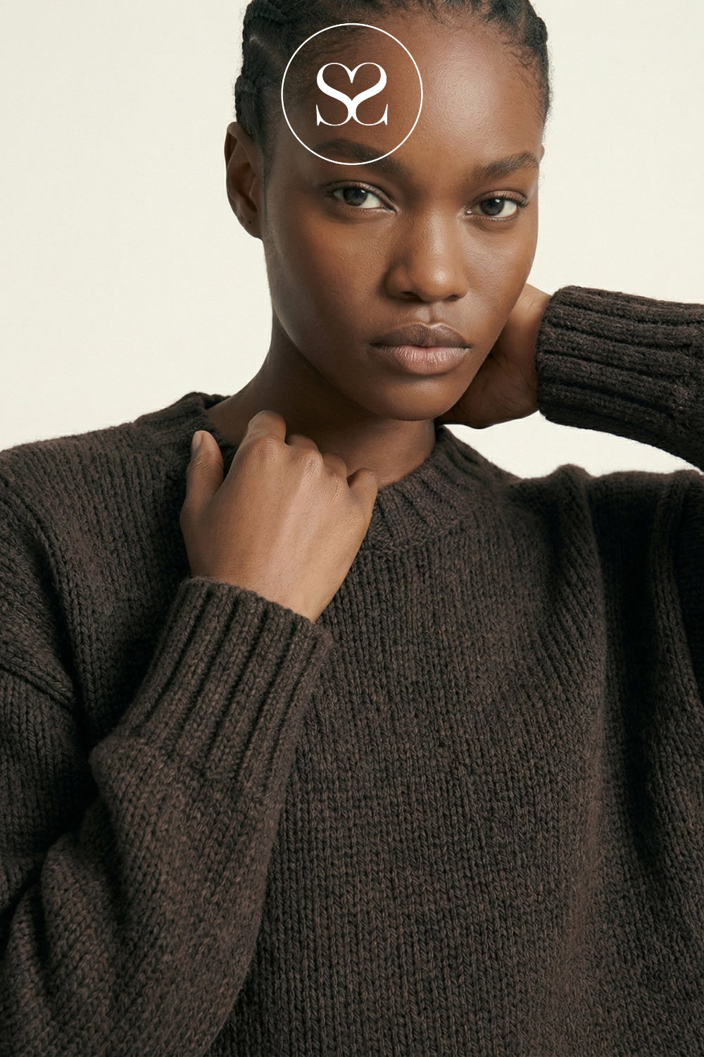 A close-up of a model wearing a dark brown ribbed knit sweater against a light beige background, with a white circular 'SS' logo overlay on her forehead.