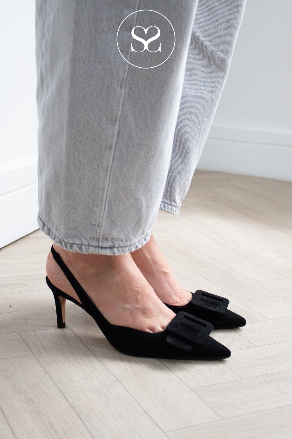 Black suede pointed-toe slingback high heels with a large square buckle detail on the toe, shown on a model wearing light gray barrel jeans on a light wood floor. Lodi Black Heels available in Ireland