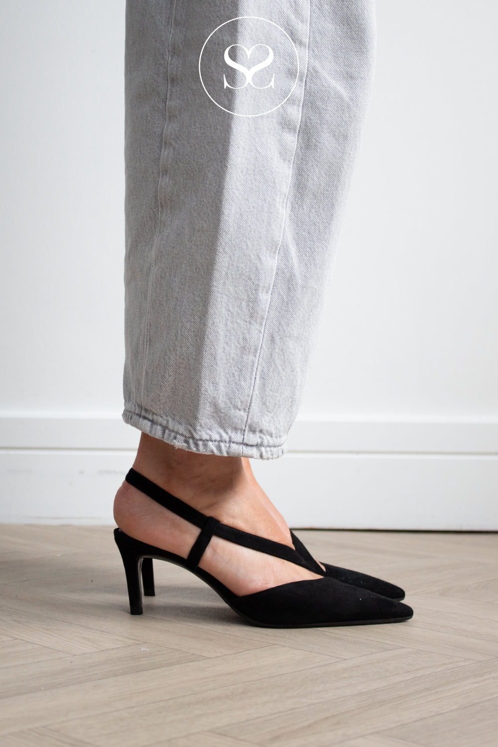 A woman wearing black slingback pointed-toe heels from Lodi with a kitten heel, paired with light gray jeans, standing on a light wood floor against a white wall.