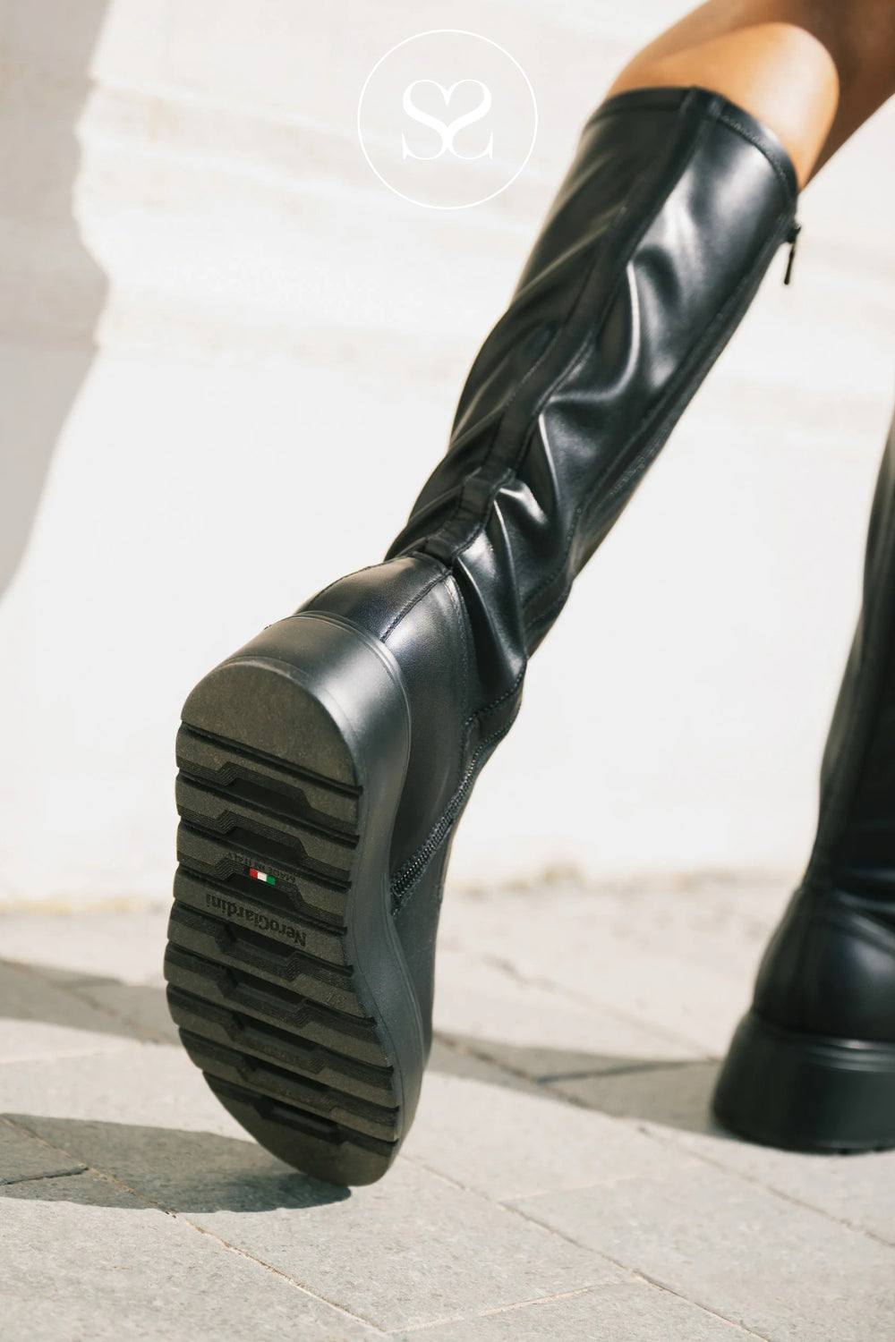 A close-up of a person wearing a tall Nero Giardini black leather knee-high boot with a chunky lug sole, shown stepping on a stone surface outdoors.