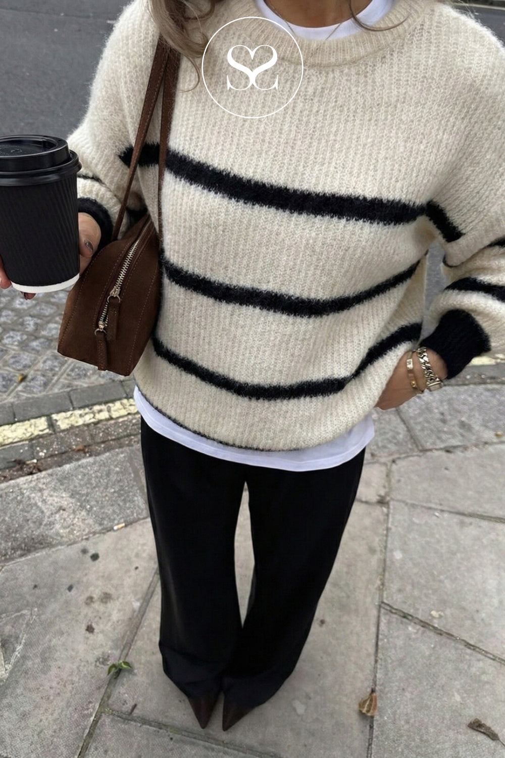 A person outdoors wearing a fuzzy cream knit sweater with bold black horizontal stripes, paired with black pants and holding a brown crossbody bag