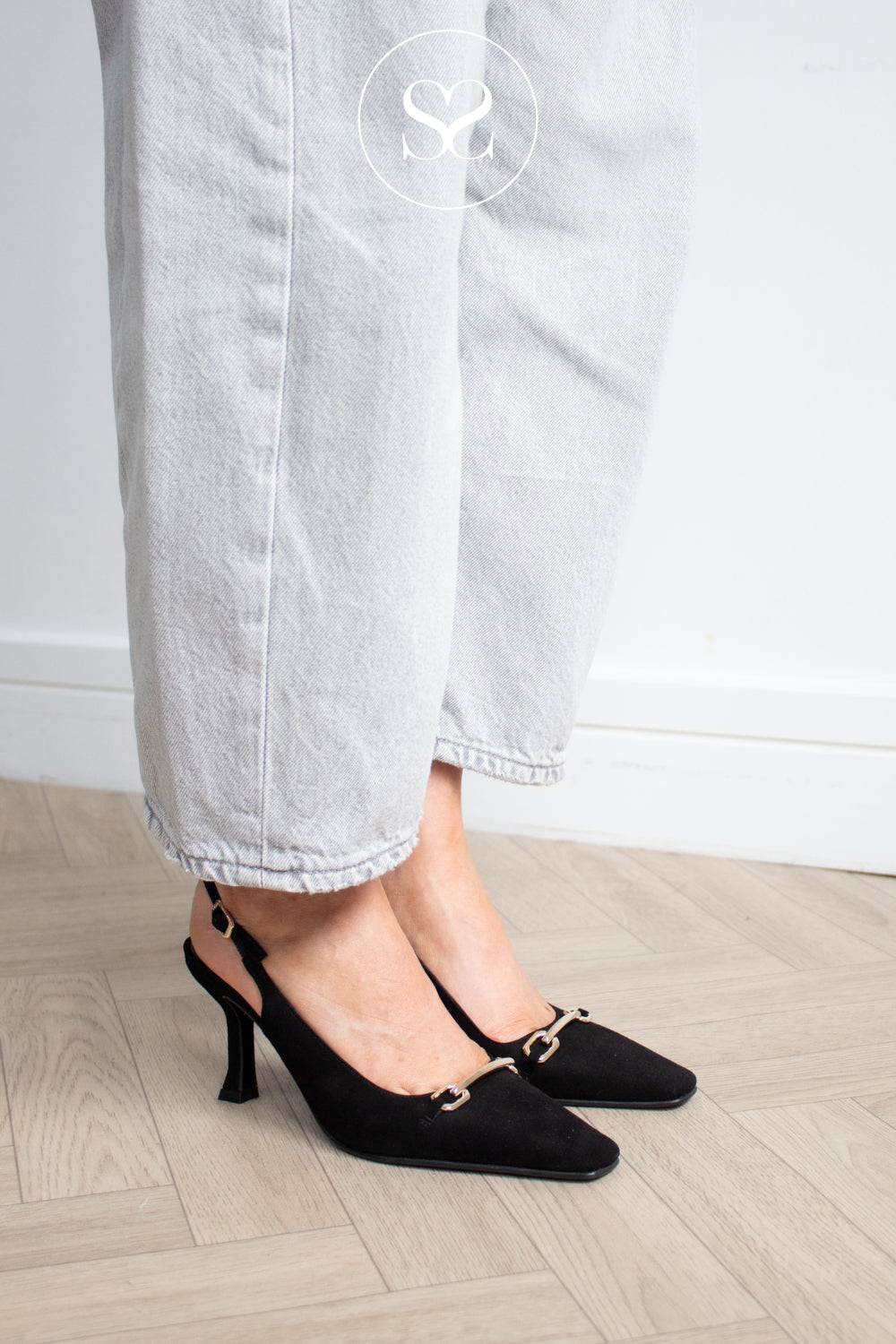 Black slingback kitten heel pumps with a small gold-tone hardware detail on the vamp, shown on a model wearing light-wash cropped jeans on a light wood floor. Lodi Black Suede Slingback High Heels - Pascal