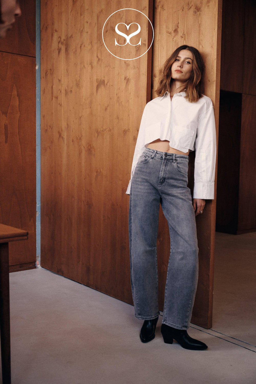 A woman stands in a doorway wearing a white cropped button-up shirt, grey barrel jeans, and black ankle boots. A circular logo with stylized letters is visible at the top center.