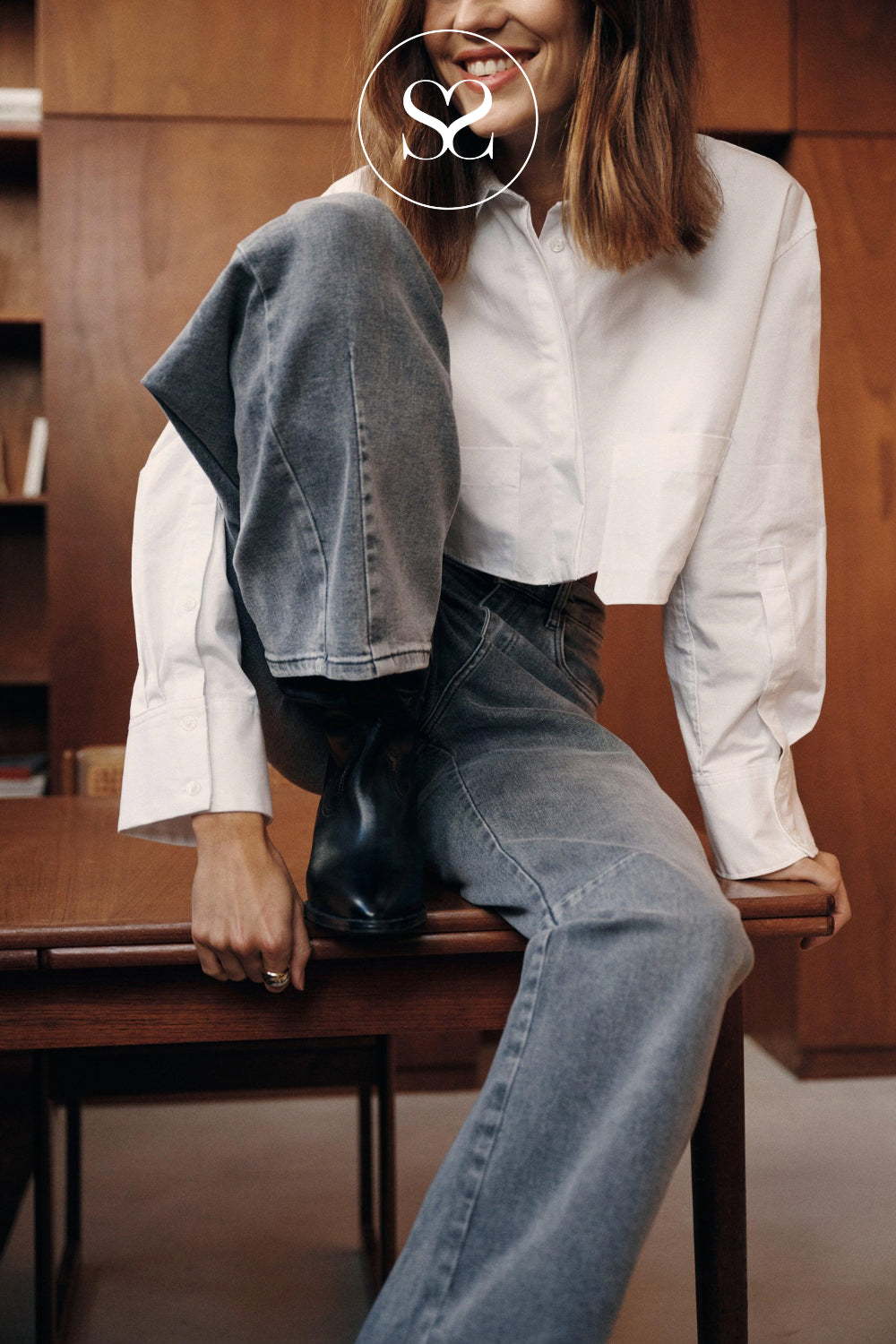 A woman sitting on a wooden table wearing a white button-up shirt, grey washed barrel jeans, and black boots stocked at sheneil shoes Ireland