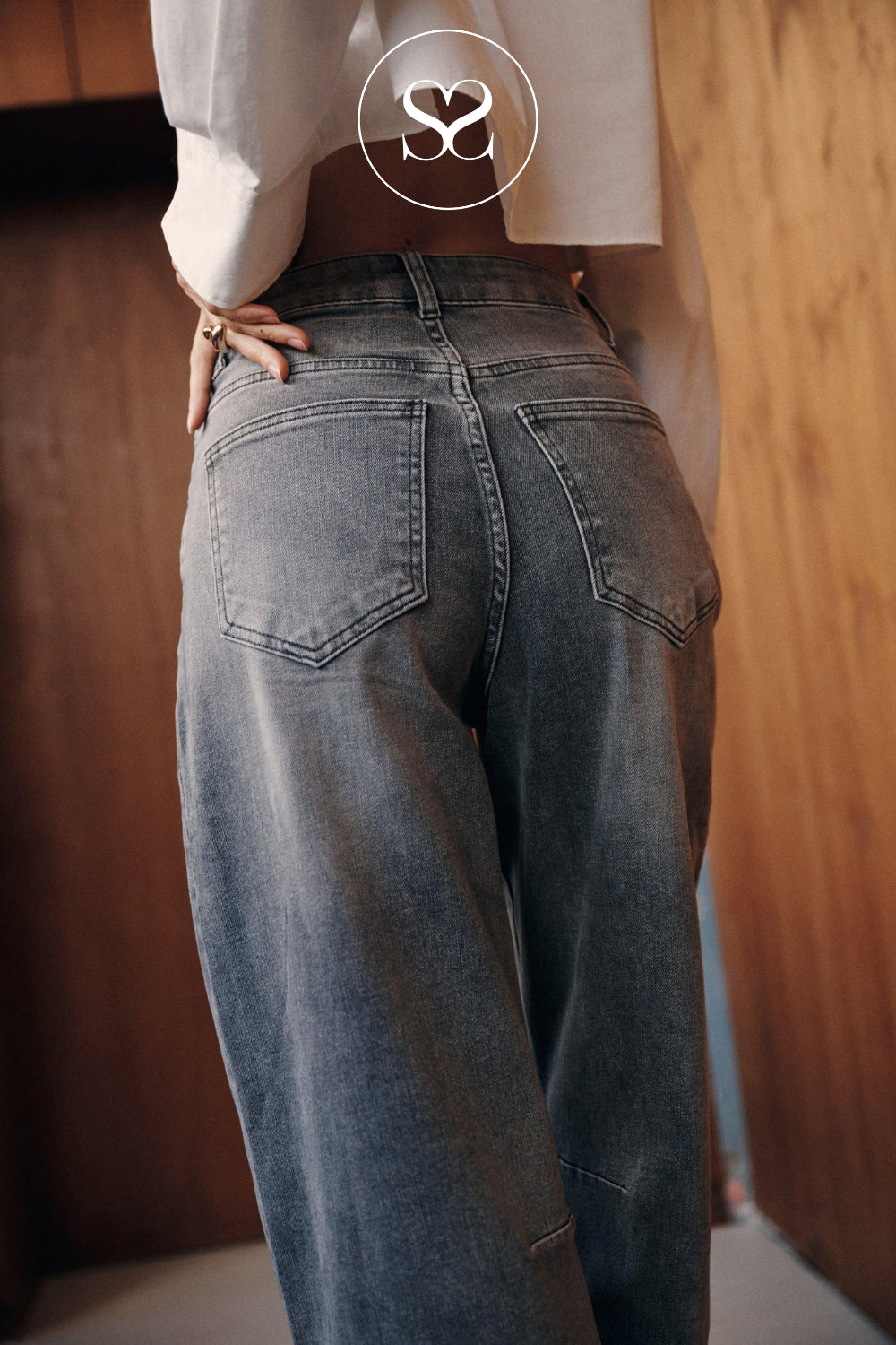 A person wearing loose barrel-leg gray denim jeans with a faded wash, shown from the back in an indoor setting with wooden walls. A white blouse is partially visible, and a circular 'SS' logo watermark is centered at the top.