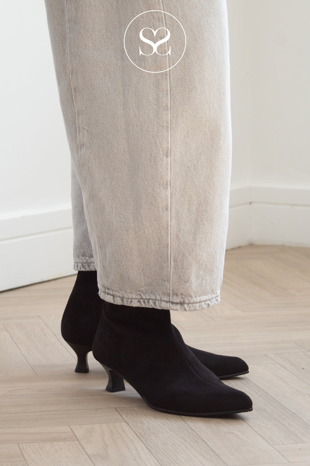 Miss Elastic Black suede pointed-toe ankle boots with kitten heels, worn with light beige wide-leg jeans on a light wood floor.