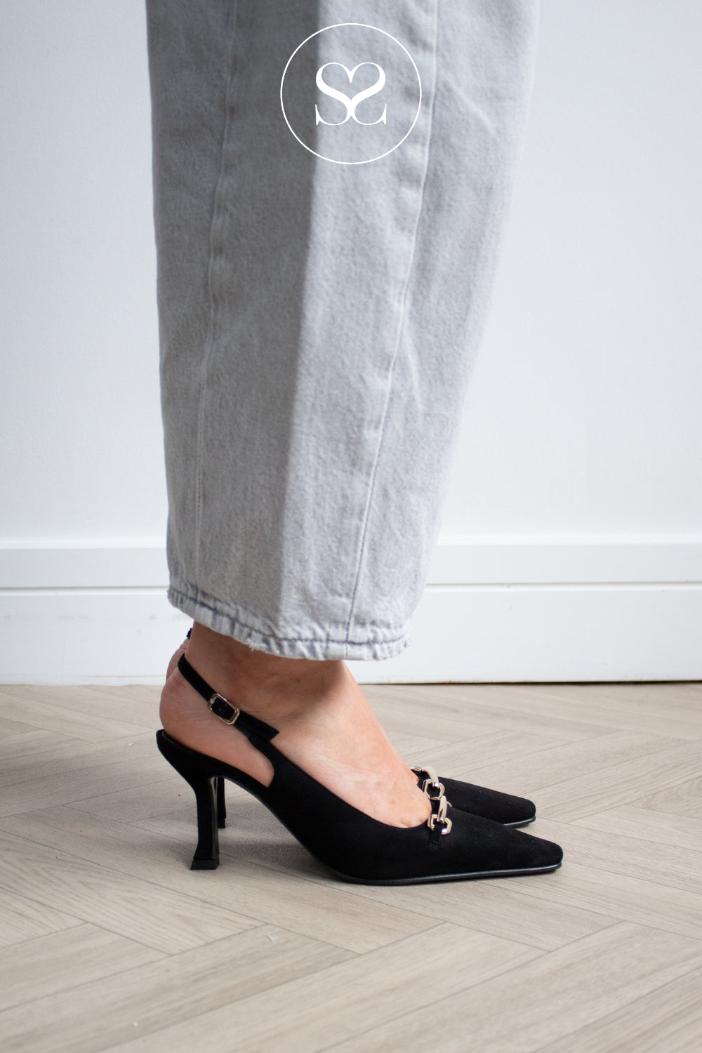 A person wearing black slingback kitten heel pumps with a small gold chain detail on the toe, paired with light grey jeans, standing on a light wood floor against a white wall. Lodi Pascal Heels in black suede