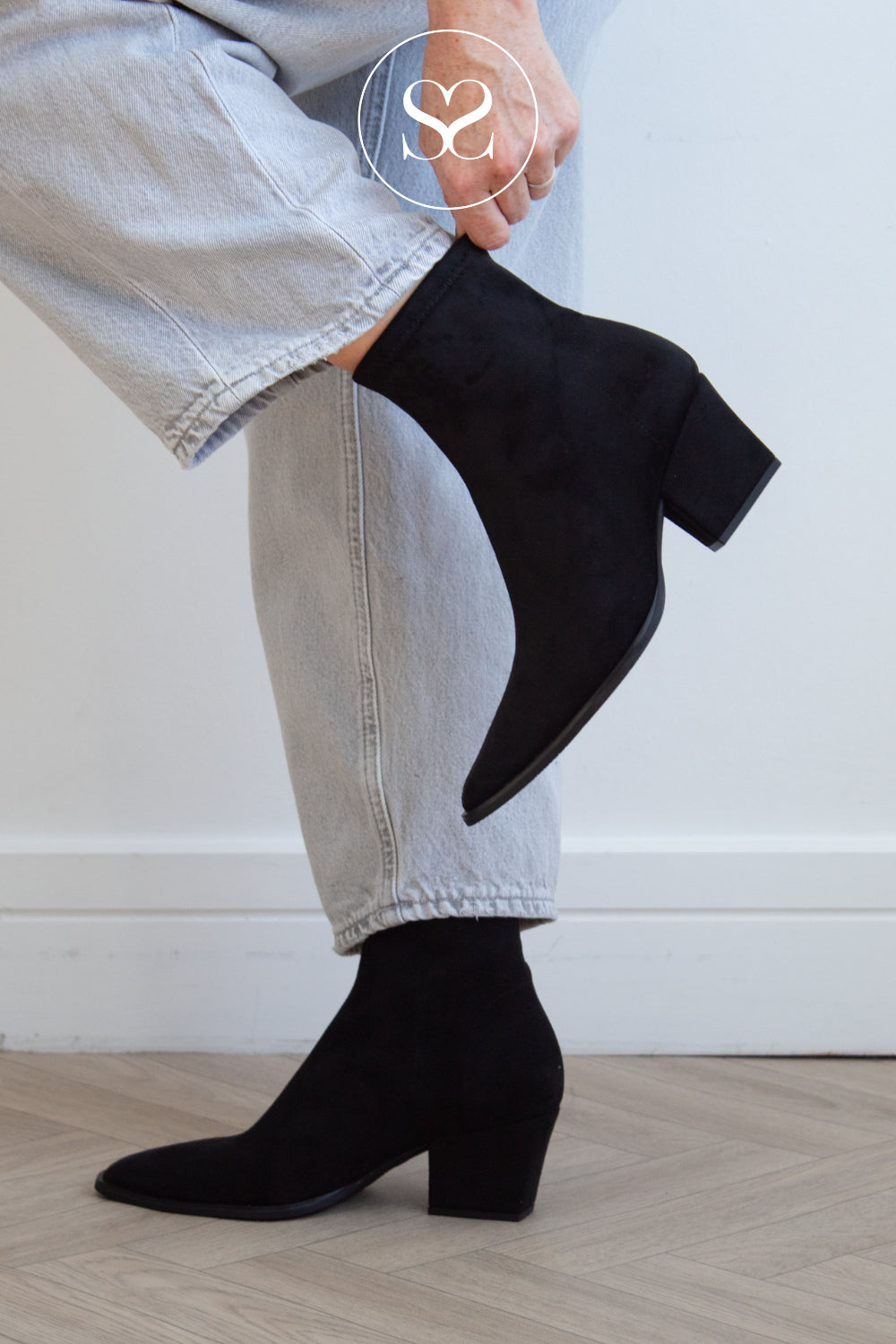 A person wearing miss elastic black suede ankle boots with pointed toes and block heels, paired with light-wash jeans, shown from the thigh down against a white wall and light wood floor.