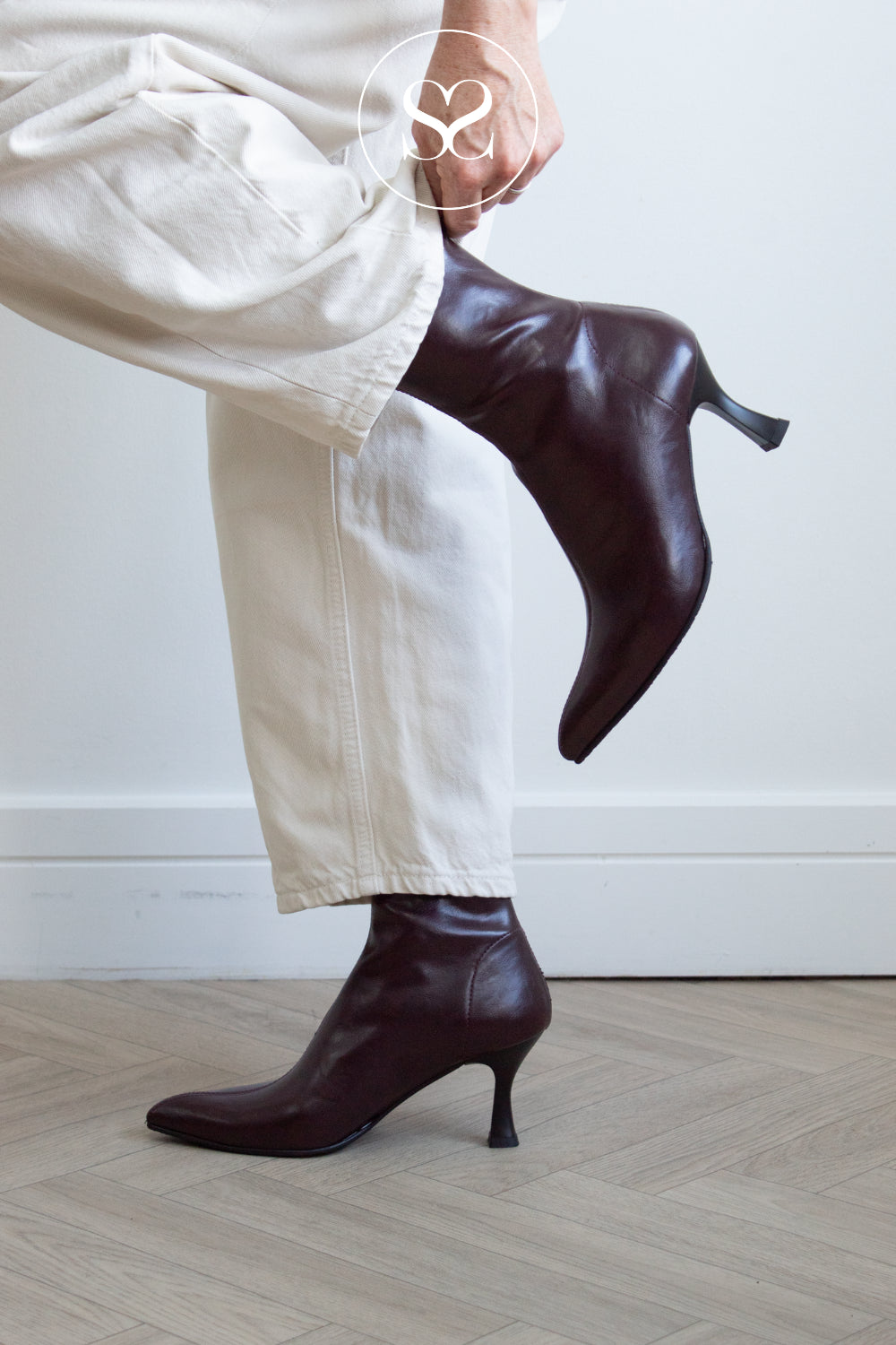 A person wearing off-white trousers and dark burgundy pointed-toe ankle boots with kitten heels from Miss Elastic, shown from the waist down against a light wall and wood floor.