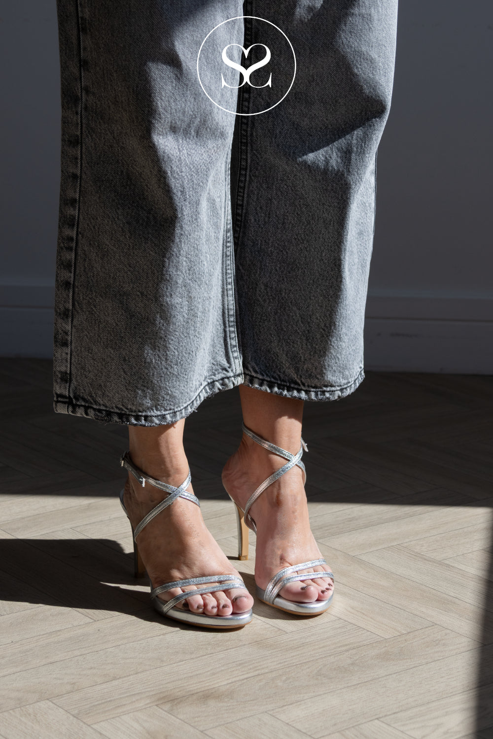 A close-up of a person's feet wearing Lodi silver metallic strappy high-heeled sandals with a slim heel, paired with wide-leg grey denim jeans. The photo is taken in a room with a light wood herringbone floor, casting shadows, and features a white circular 'SS' logo watermark at the top.
