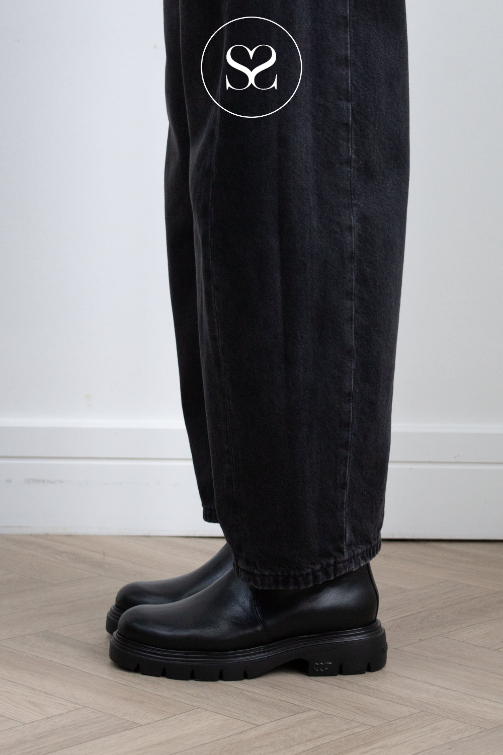 Woman wearing Wonders Black leather Chelsea boots with chunky lug soles worn with black wide-leg jeans, shown from the knees down on a light wood floor against a white wall. Shot in Ireland