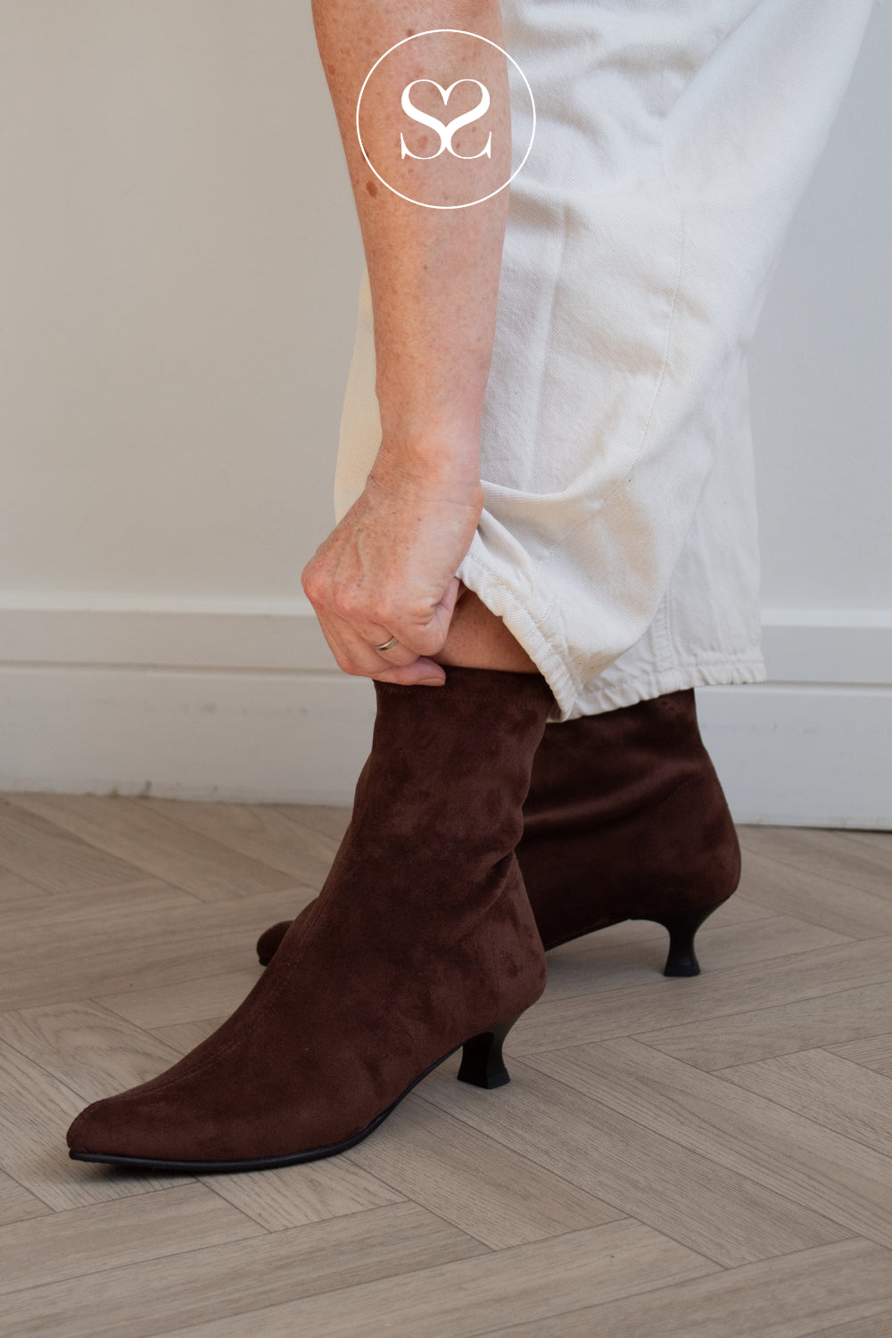 A Woman wearing Miss Elastic dark brown suede ankle boots with kitten heels and pointed toes, paired with white trousers, standing on a light wood herringbone floor. From Sheneil Shoes Galway Ireland