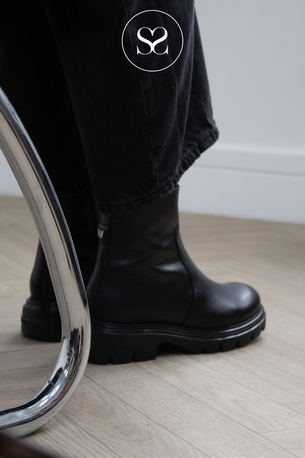 A close-up of a woman wearing Wonders black leather ankle boots with black jeans, standing on a light wood floor next to a reflective silver metal chair leg. A circular logo with a Sheneil Shoes logo inside a circle is visible near the top.